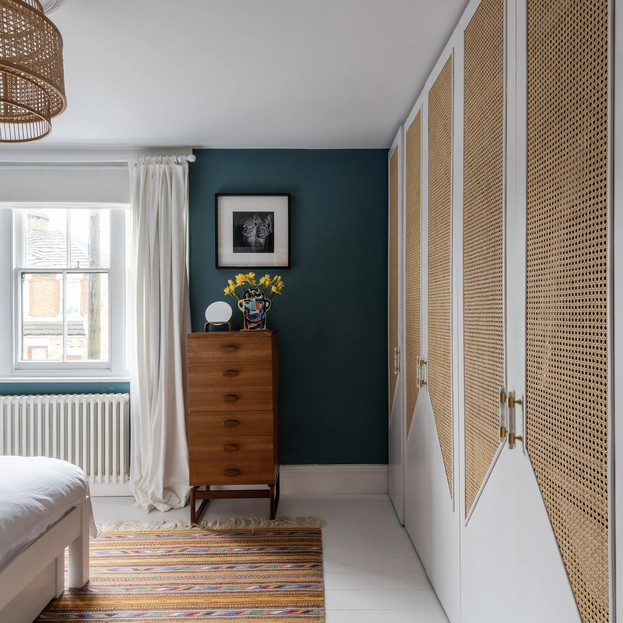 A bedroom corner with a teal wall, a window with white curtains, a striped rug, a wooden dresser with a vase of yellow flowers and a small lamp, a framed black-and-white photograph above, and white closet doors with rattan panels.