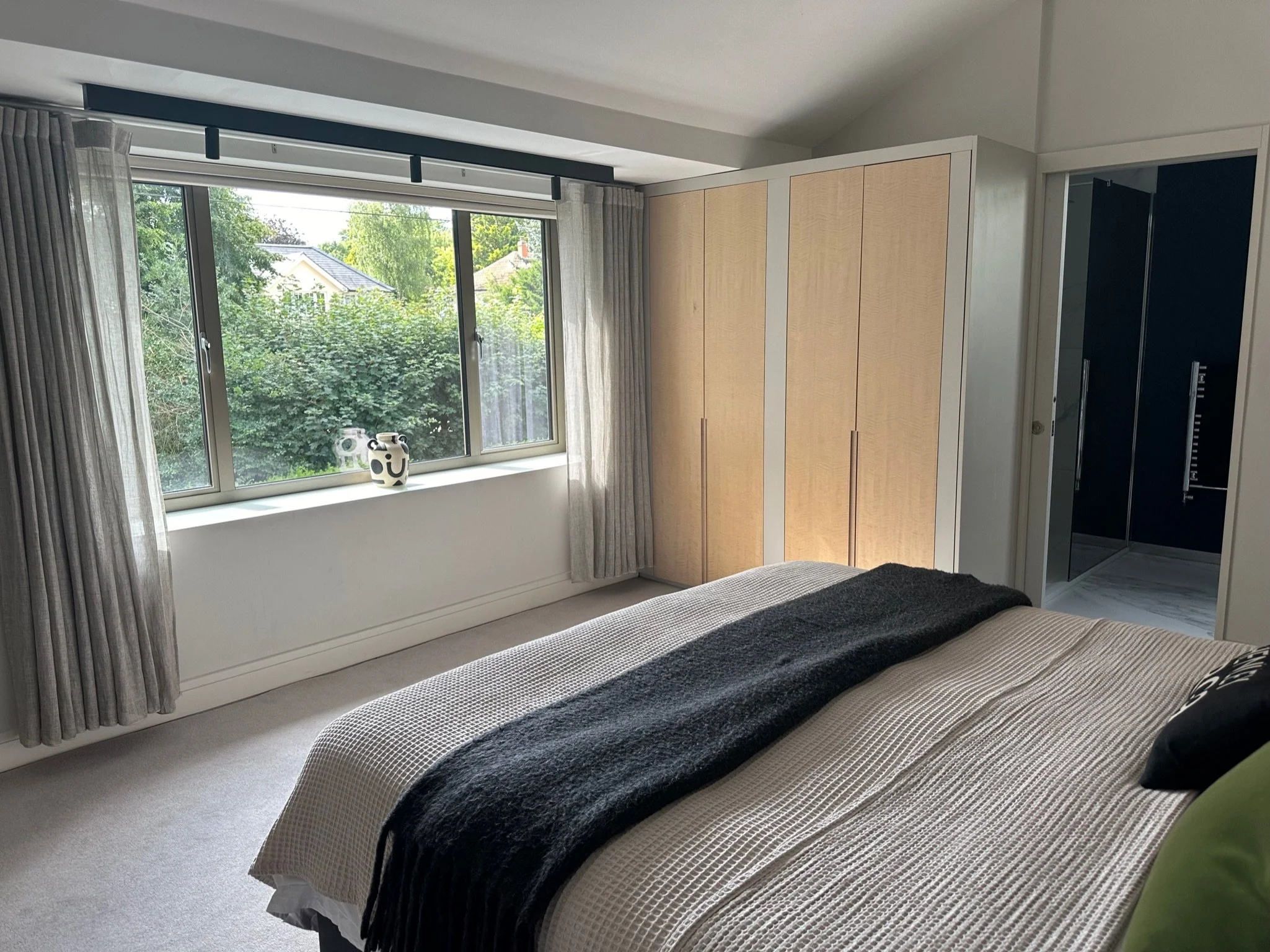A bedroom with a large window dressed with beige curtains, a white windowsill with two decorative vases, a bed with beige and black bedding, a wooden wardrobe, and a doorway leading to a bathroom with black walls and a shower.