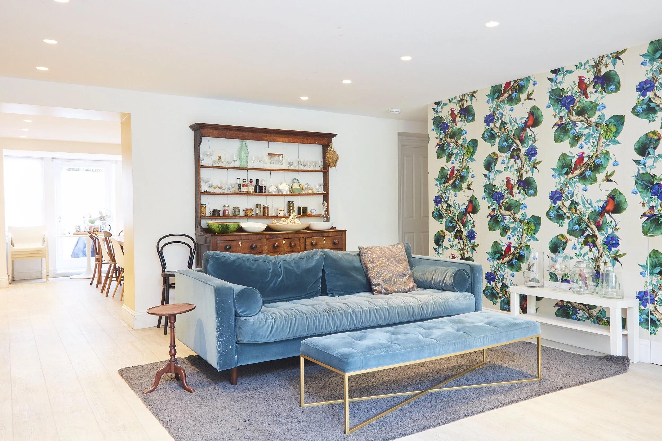 Living room with a blue velvet sofa, tufted ottoman, and a patterned throw pillow. A white side table with glass vases is nearby. A dark wood hutch displays glassware and decorative items. The wall behind the sofa features colorful tropical bird and 