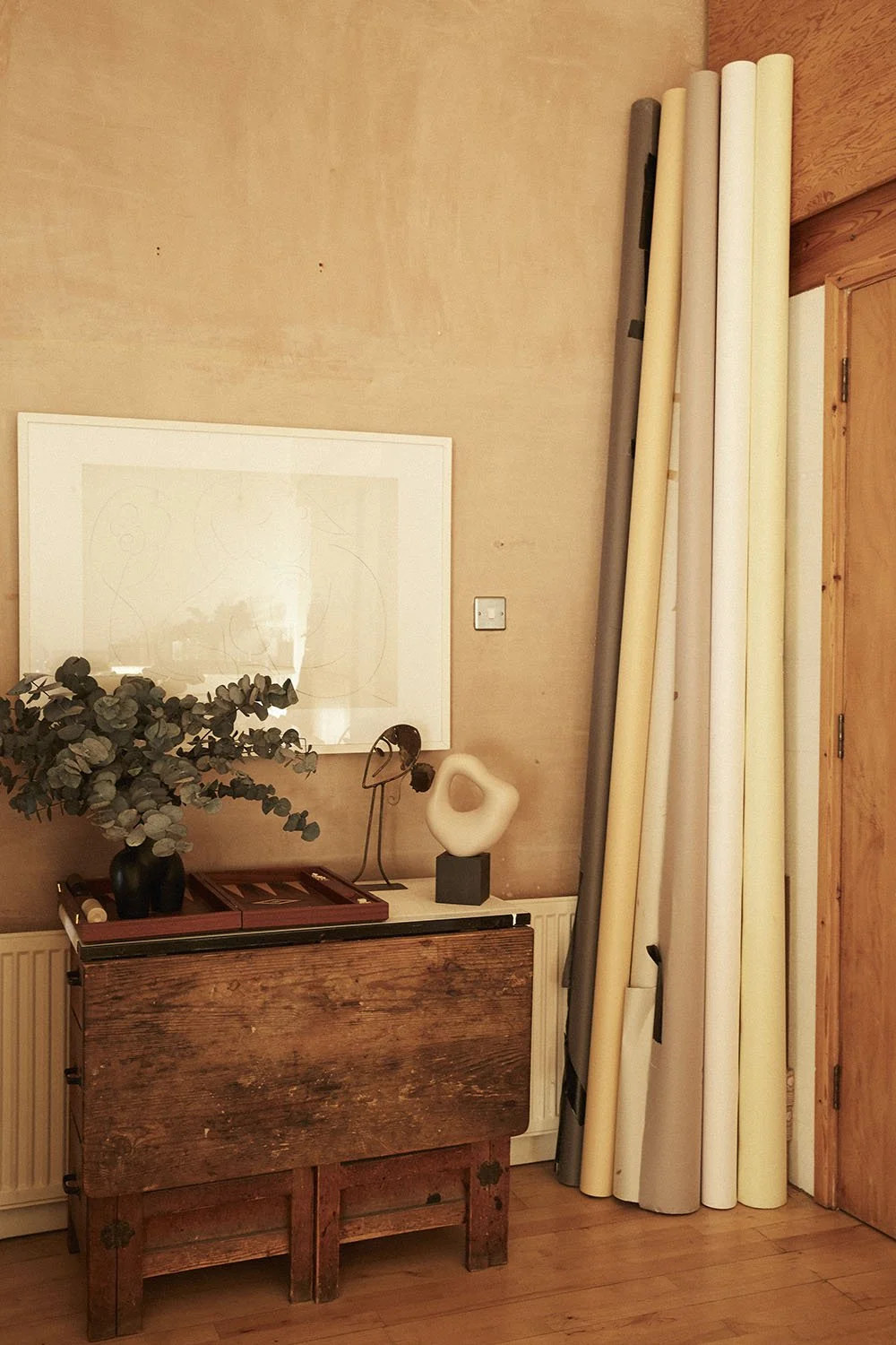A wooden sideboard with a potted eucalyptus plant, a modern sculpture, and a small tray with a candle. Several rolled-up posters or canvases are leaning against the wall next to a wooden door.