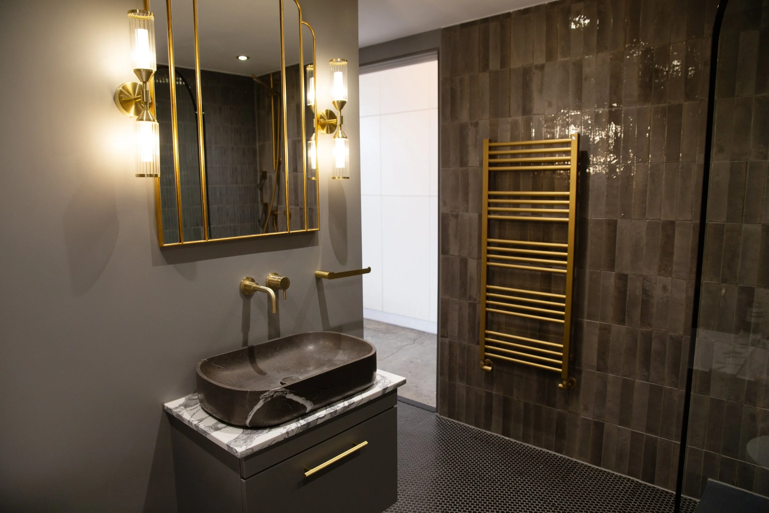 Modern bathroom with dark tiled wall, gold towel radiator, marble countertop with a dark sink, and gold fixtures, illuminated by wall-mounted lights and natural light from a large window.