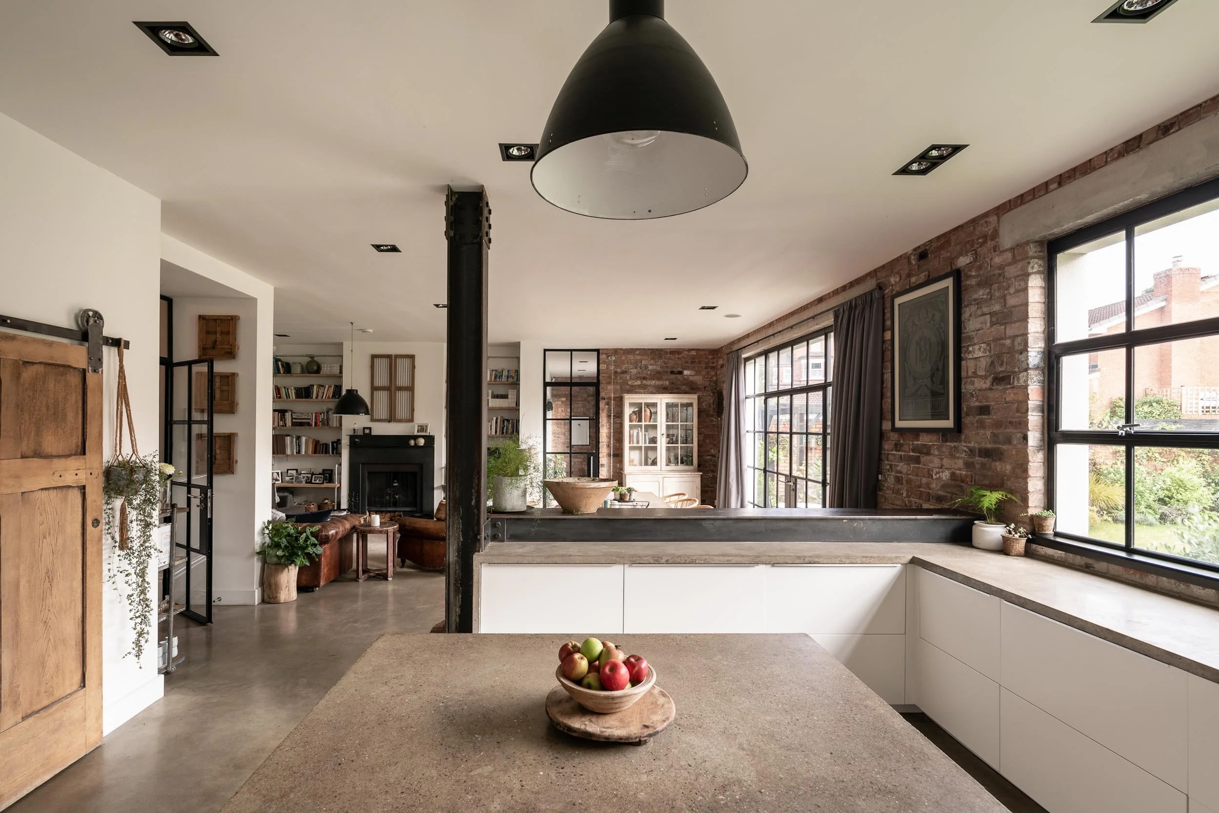 Modern open-plan kitchen and living room with large windows, brick wall, white cabinets, wooden accents, and decorative plants.