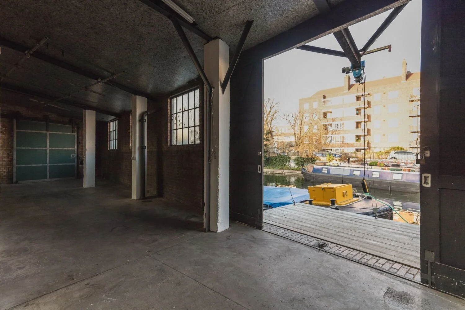 Empty indoor garage with brick walls, chipped paint, and a large open doorway leading to a docked boat outside on a canal.