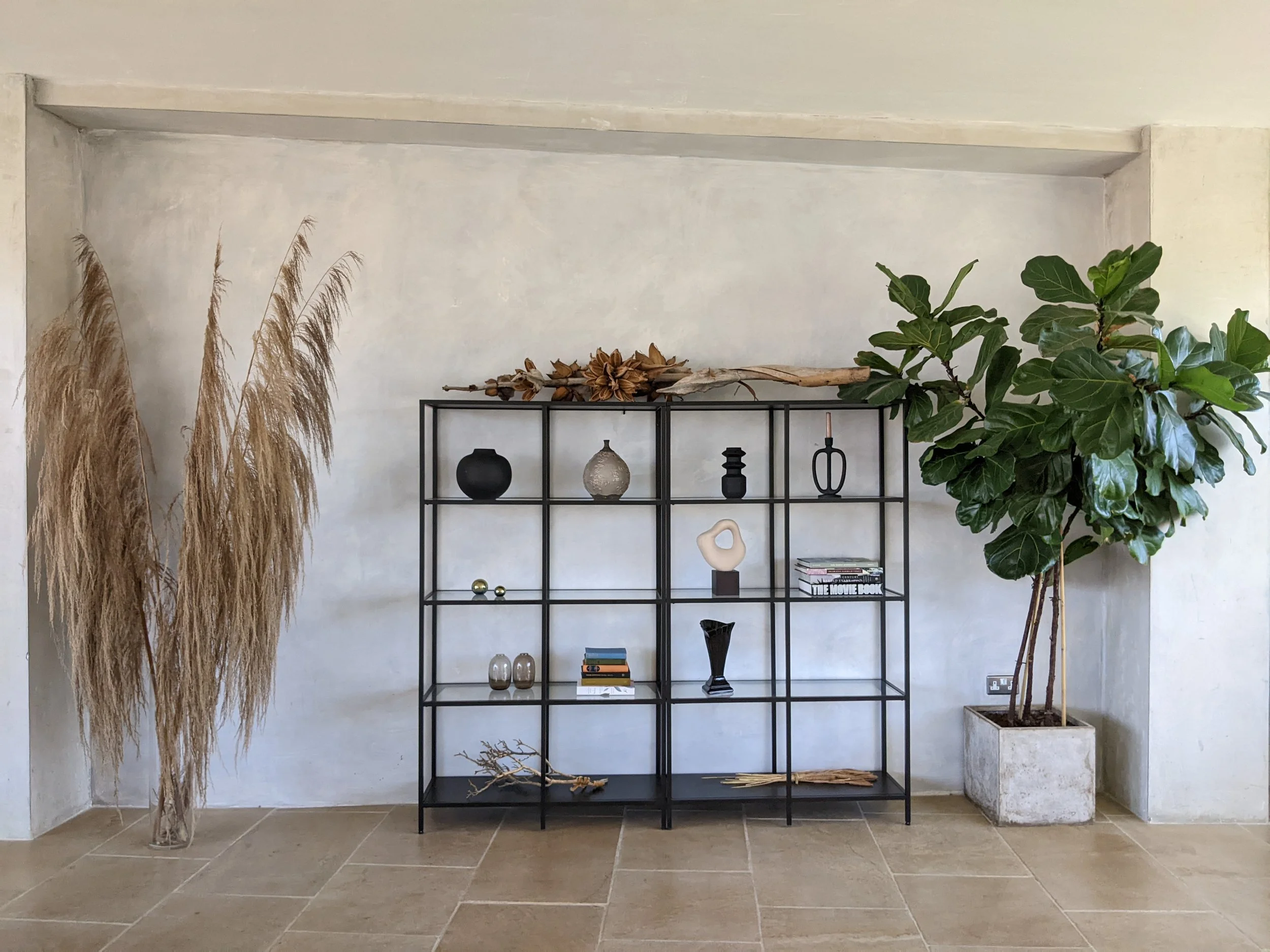 Decorative black metal shelf with various vases and books, tall beige pampas grass on the left, large green leafy plant in a concrete planter on the right, on a beige tiled floor against a light-colored wall.