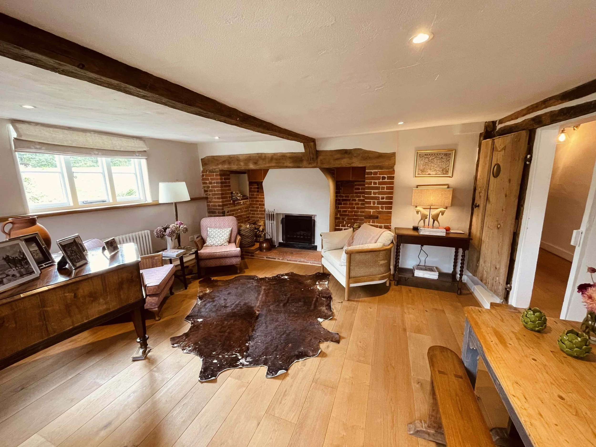 Living room with brick fireplace, pink chairs, wooden beams, large window, table with picture frames, lamps, and a cowhide rug.