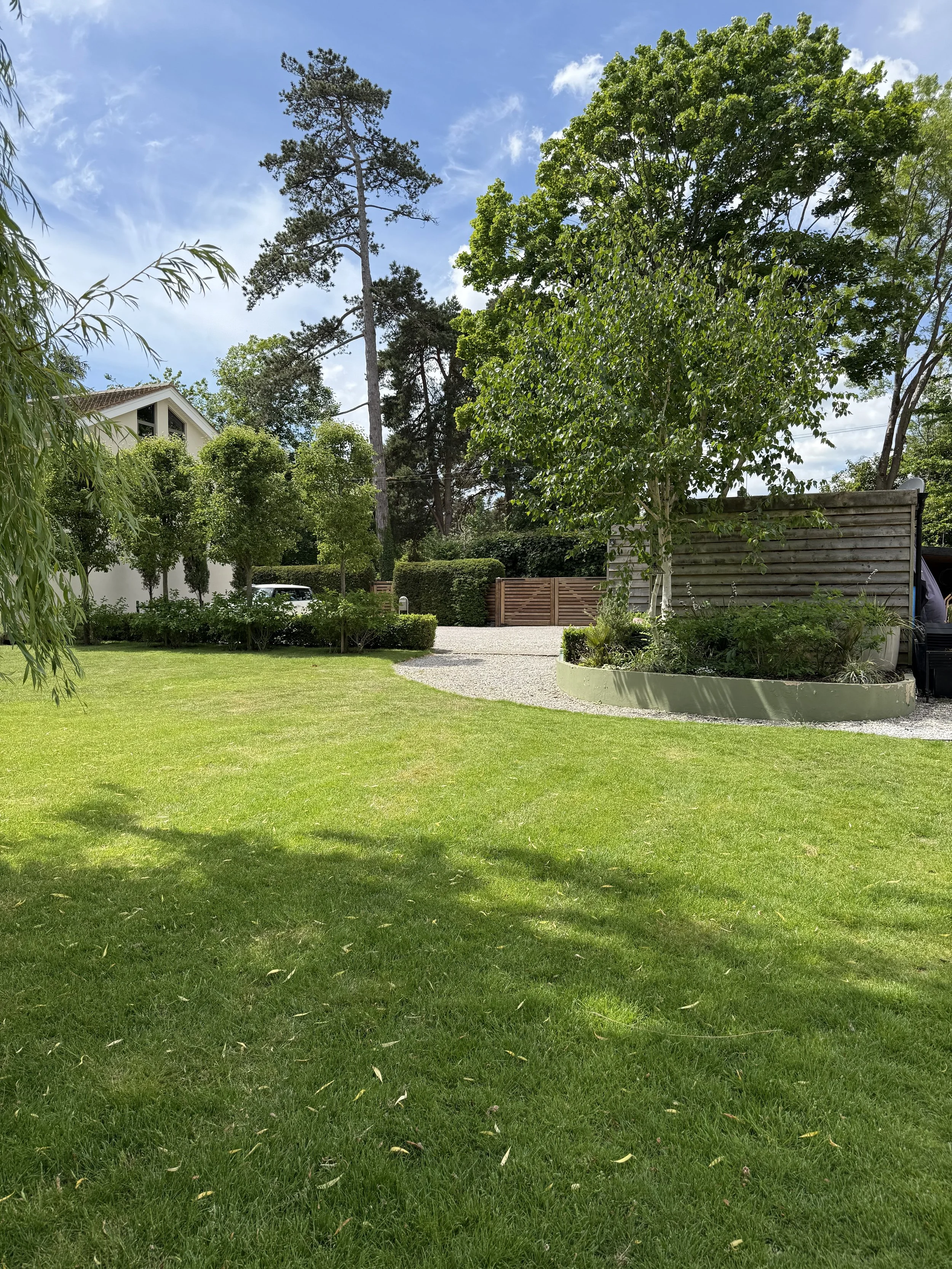 A lush green backyard with well-maintained grass, trees, and shrubs, a gravel pathway, a wooden fence, and a modern house partially visible on the left. There's also a wooden privacy screen and a parked vehicle in the distance, all under a partly clo