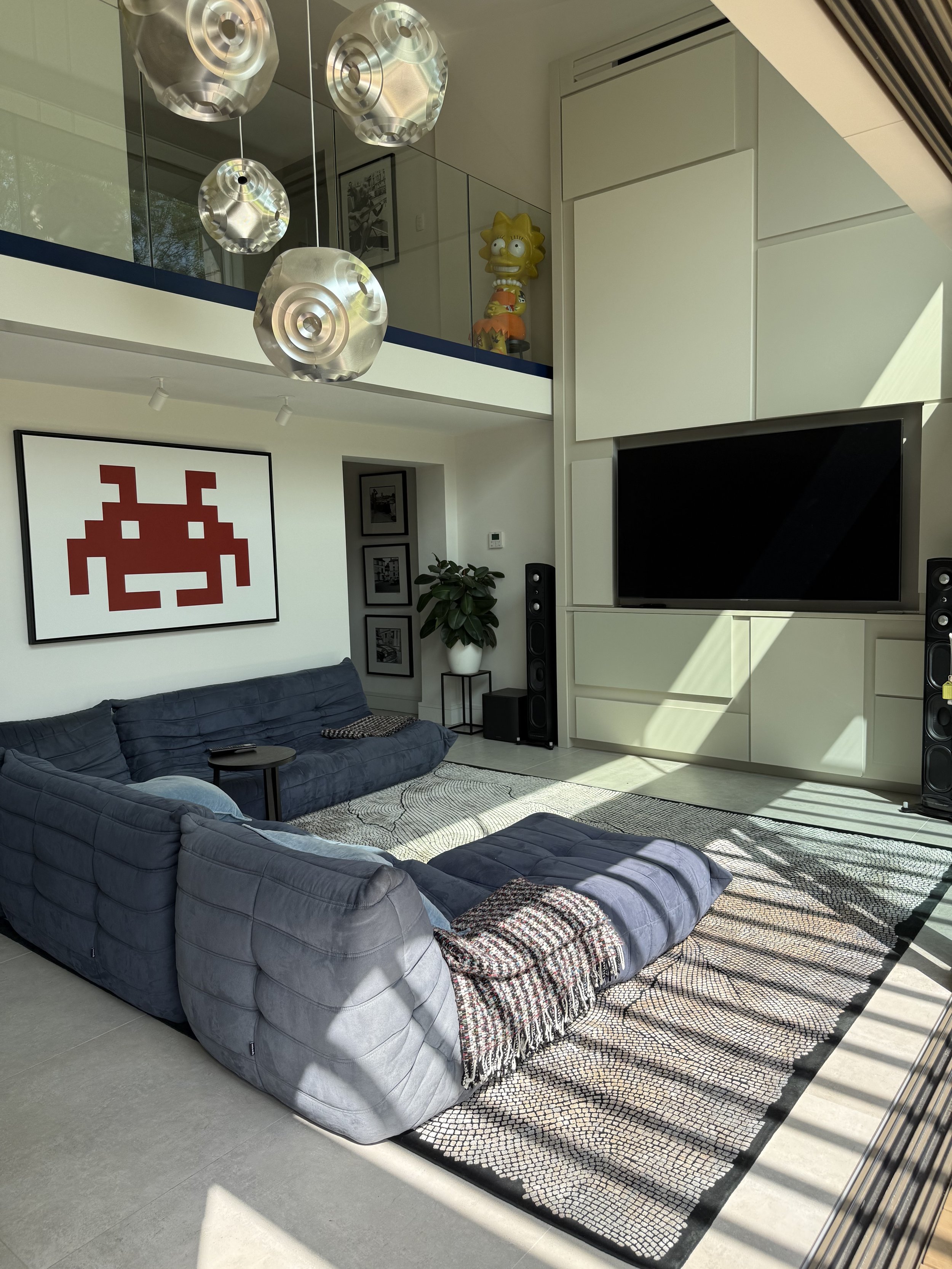 Modern living room with a large flat-screen TV, blue sectional sofa with throw blankets, a geometric patterned area rug, a potted plant, wall art including a pixel art of a red alien, and decorative pendant lights hanging from the ceiling. A Lisa Sim
