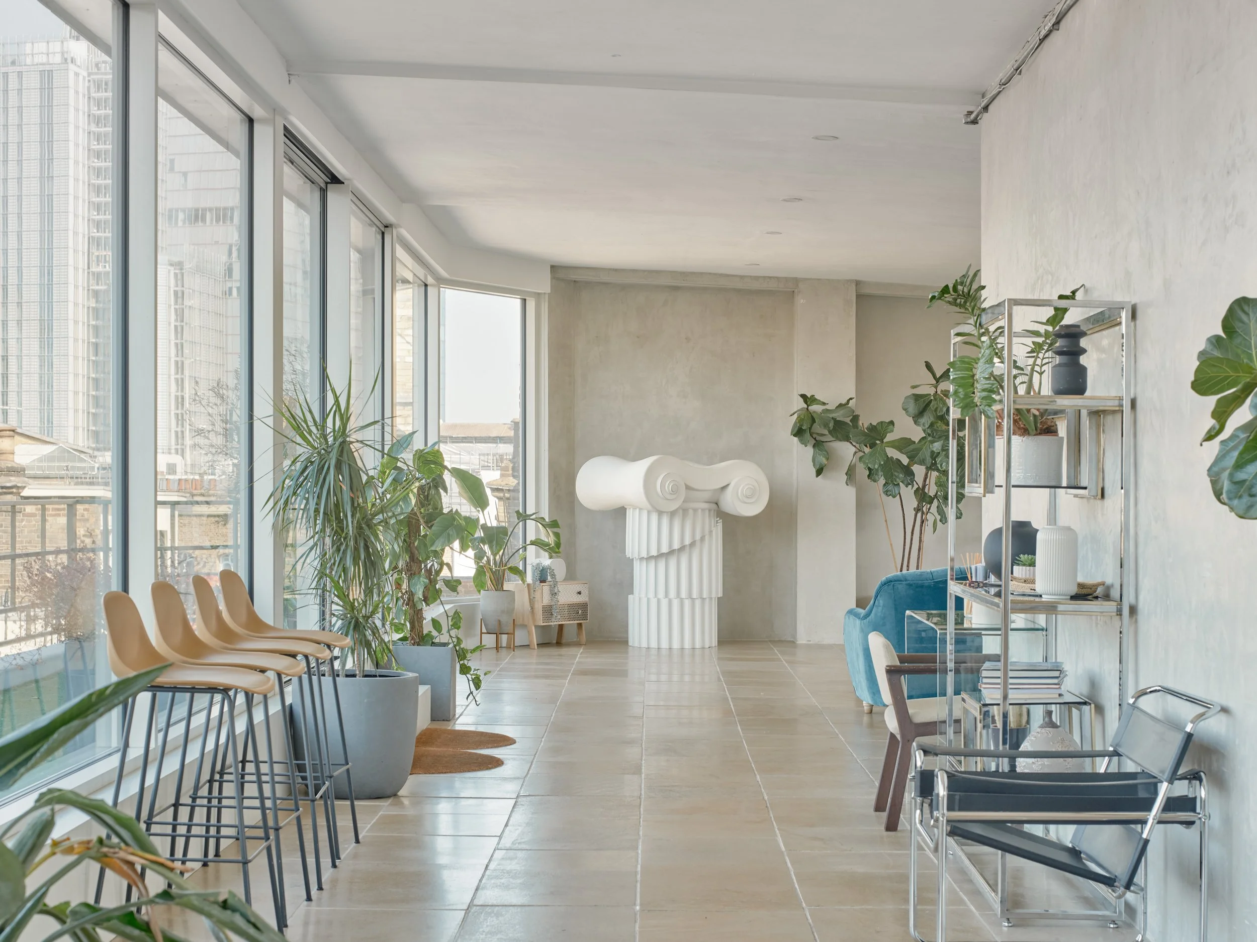 Modern interior space with large windows, potted plants, beige chairs on the left, a blue sofa, shelving unit with decorative items, and a sculptural column with spiral design.
