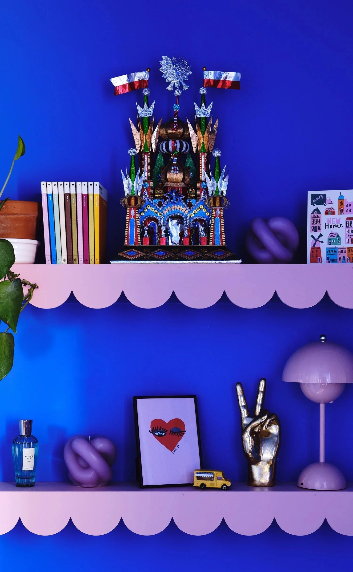 Interior of a room with two pink scalloped shelves against a blue wall. The top shelf has a colorful decorative castle figurine with two small American flags, a white decorative snowflake, purple pumpkin-shaped objects, and a colorful card. The botto
