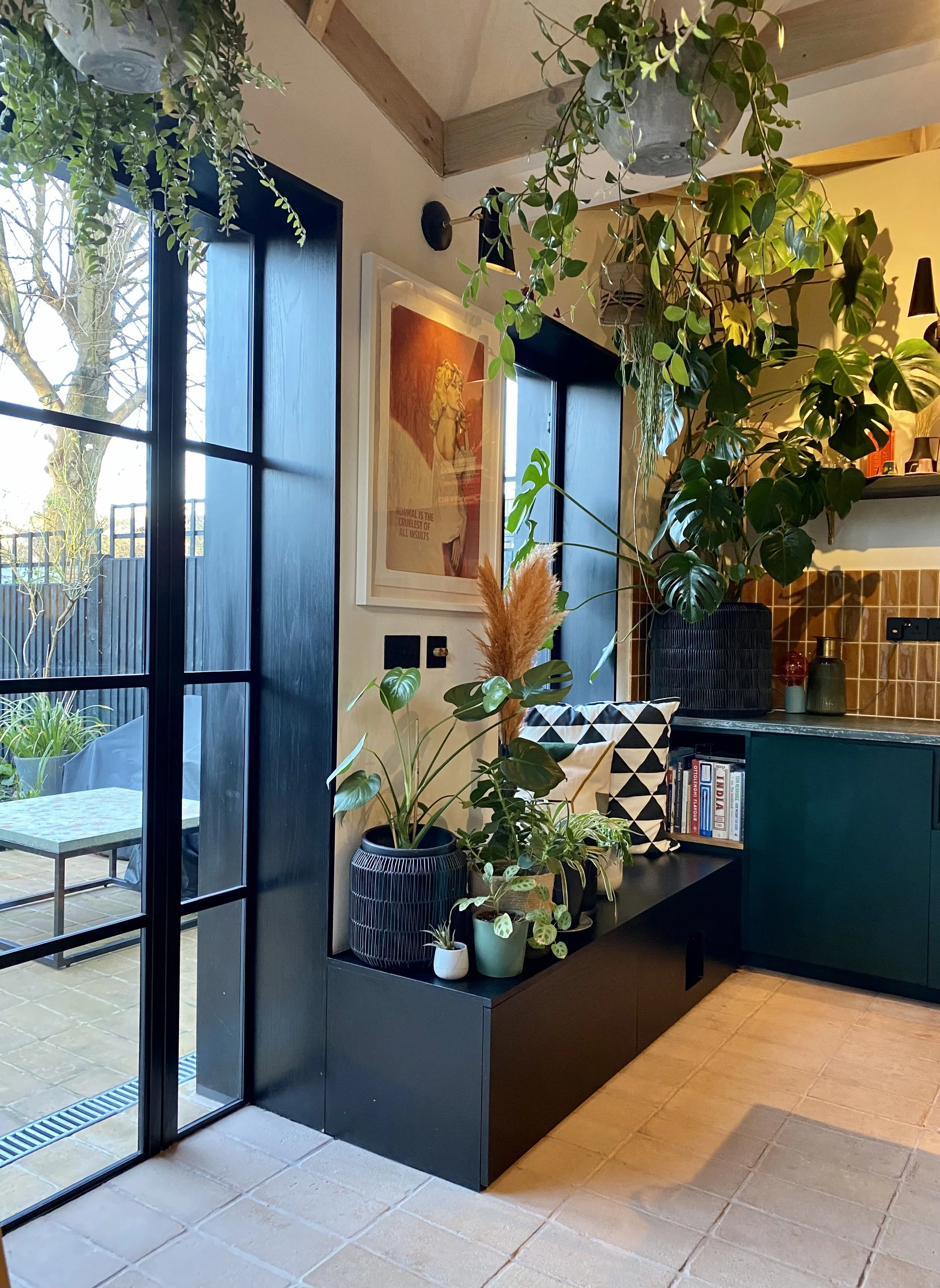 Interior view of a cozy room with large, leafy green plants in decorative pots, a framed art print on the wall, a black cabinet with books, and a large window opening to an outdoor patio with trees and a table.