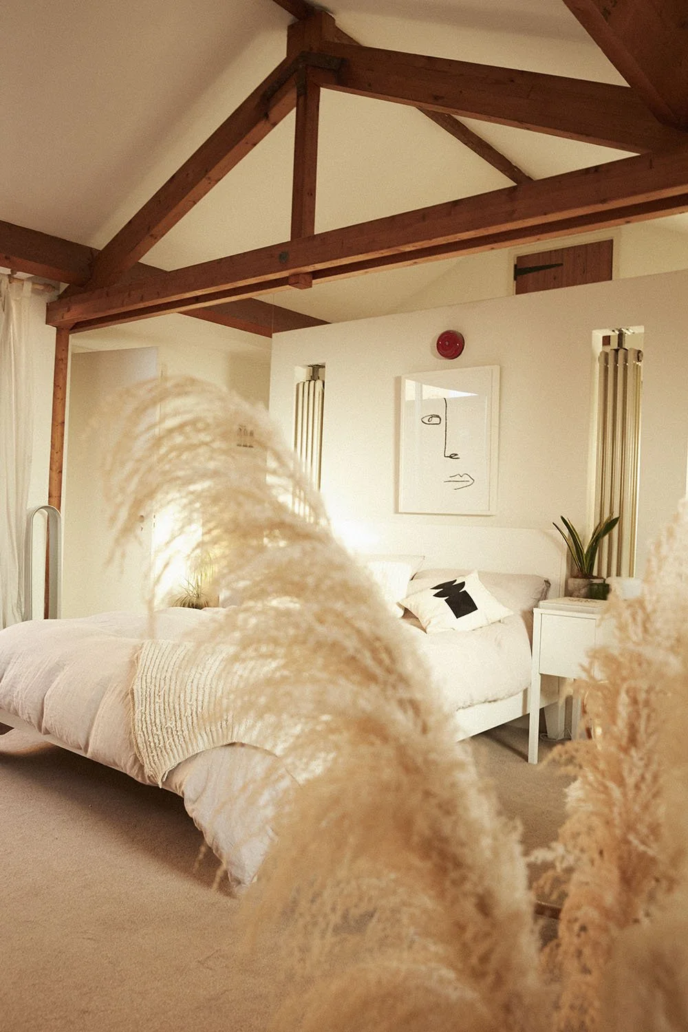 A cozy bedroom with a white bed, minimalist artwork, and a bedside table with a plant. Exposed wooden beams are visible on the ceiling, and a curtain is partially visible on the left. The photo is taken from a low angle with some dried plants in the 