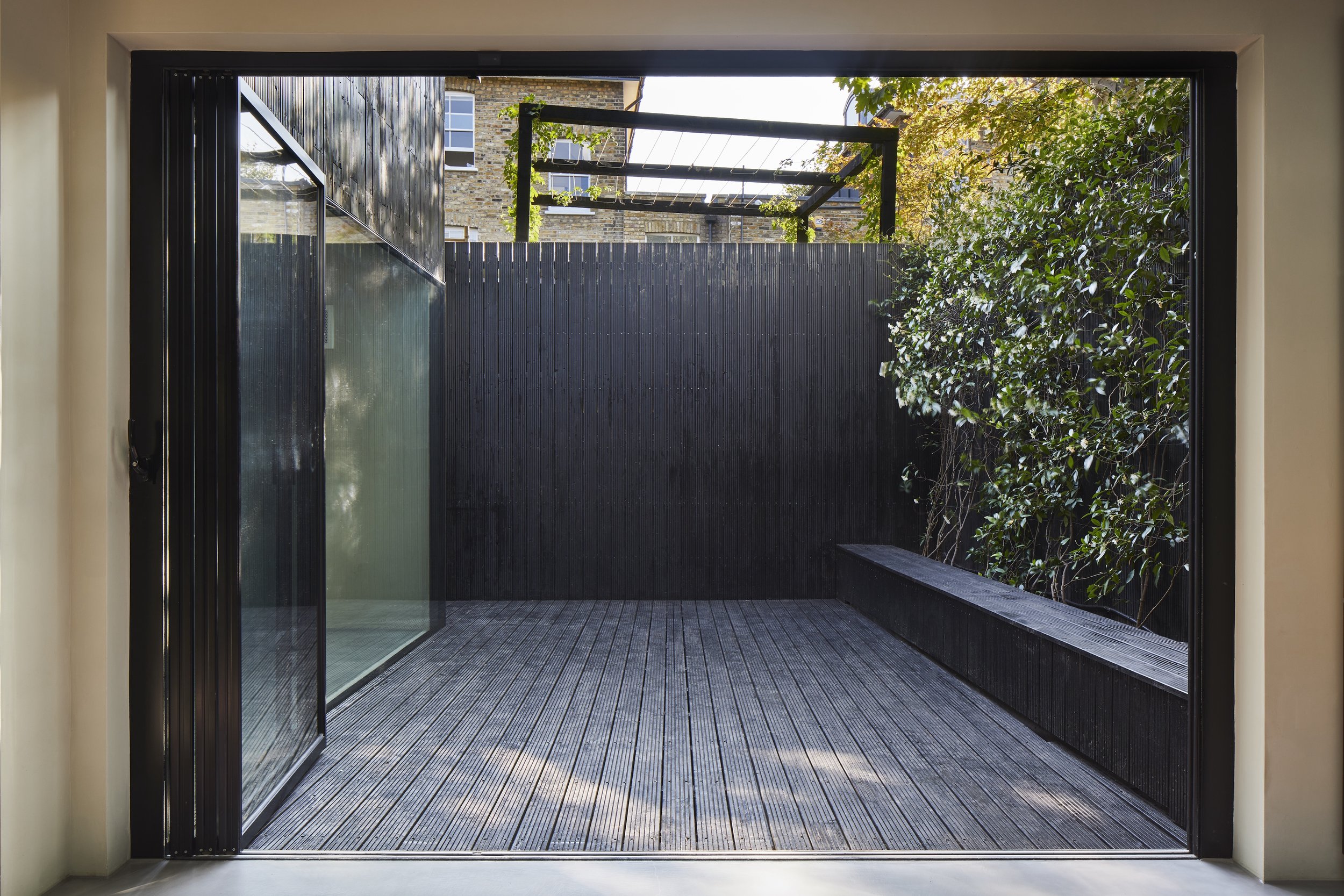 A small outdoor patio with wooden decking, black fence, and a built-in bench on the right side. There are glass sliding doors on the left leading inside. A tree with green leaves is visible on the right, and a brick building is seen in the background