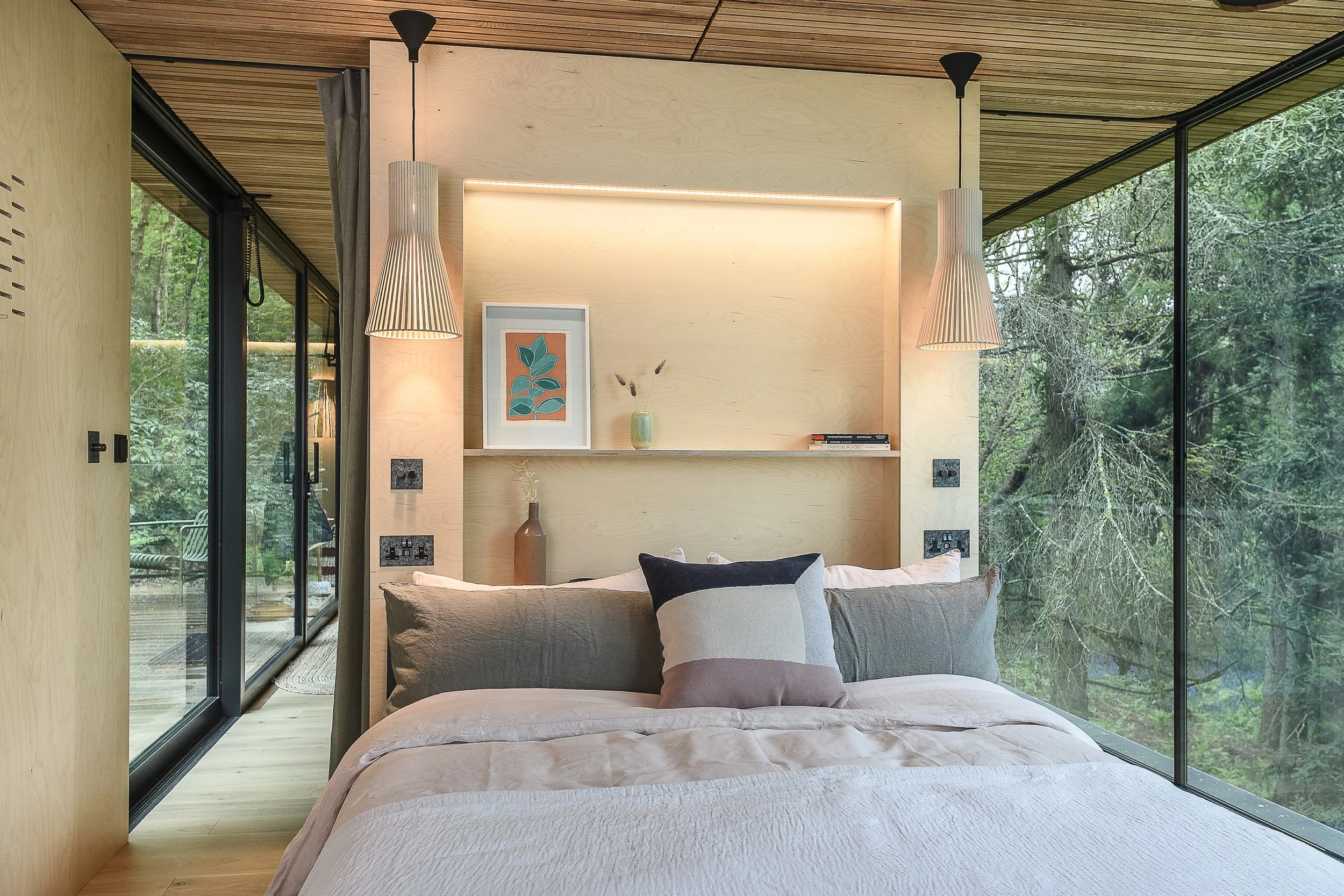 Bedroom with large glass windows showing lush trees outside, wooden ceiling, modern hanging lights, light wood headboard, framed artwork, decorative vases, and pillows on the bed.