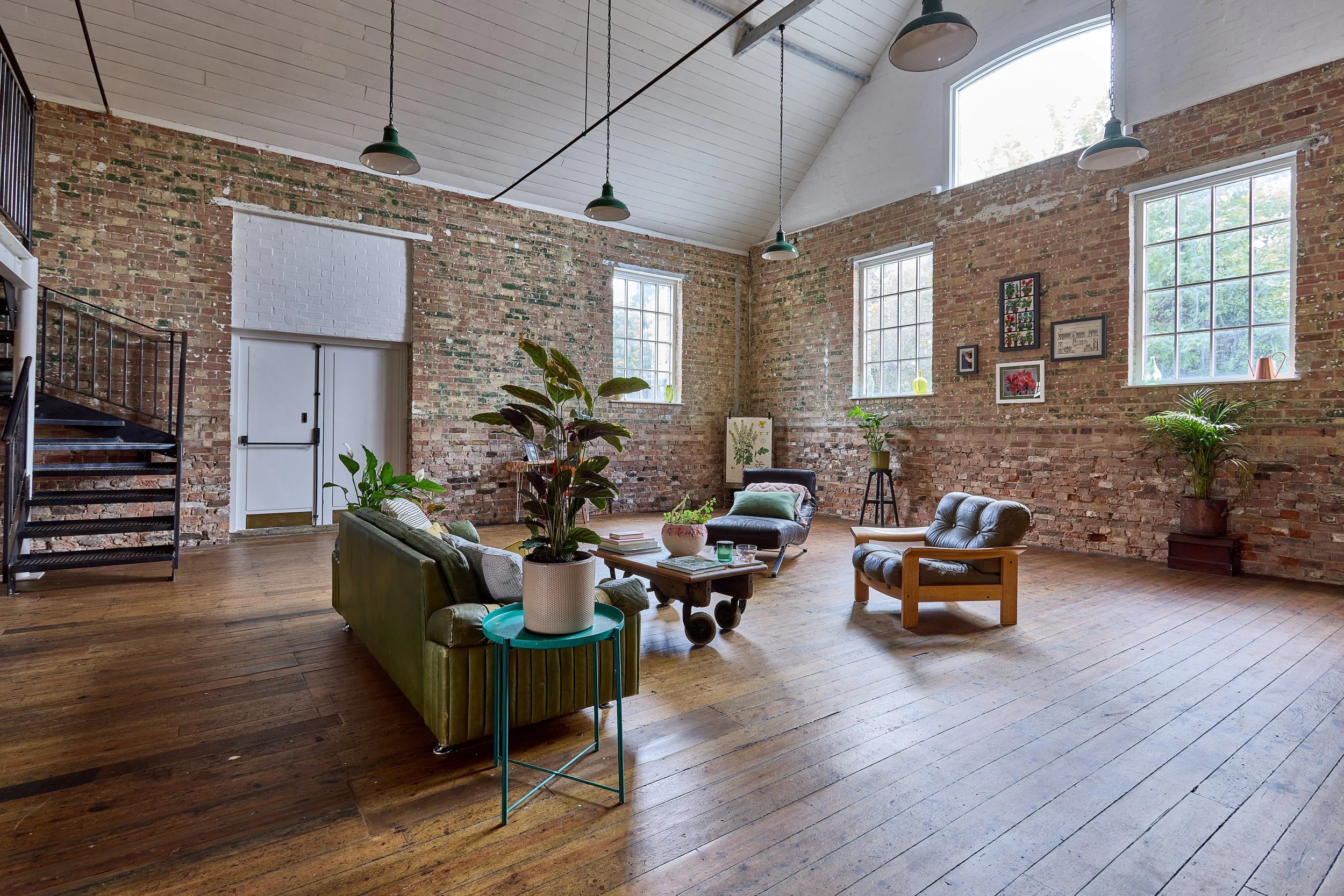 An industrial-style living room with exposed brick walls, large windows, and wooden flooring. The room contains vintage and modern furniture including sofas, chairs, and plants.