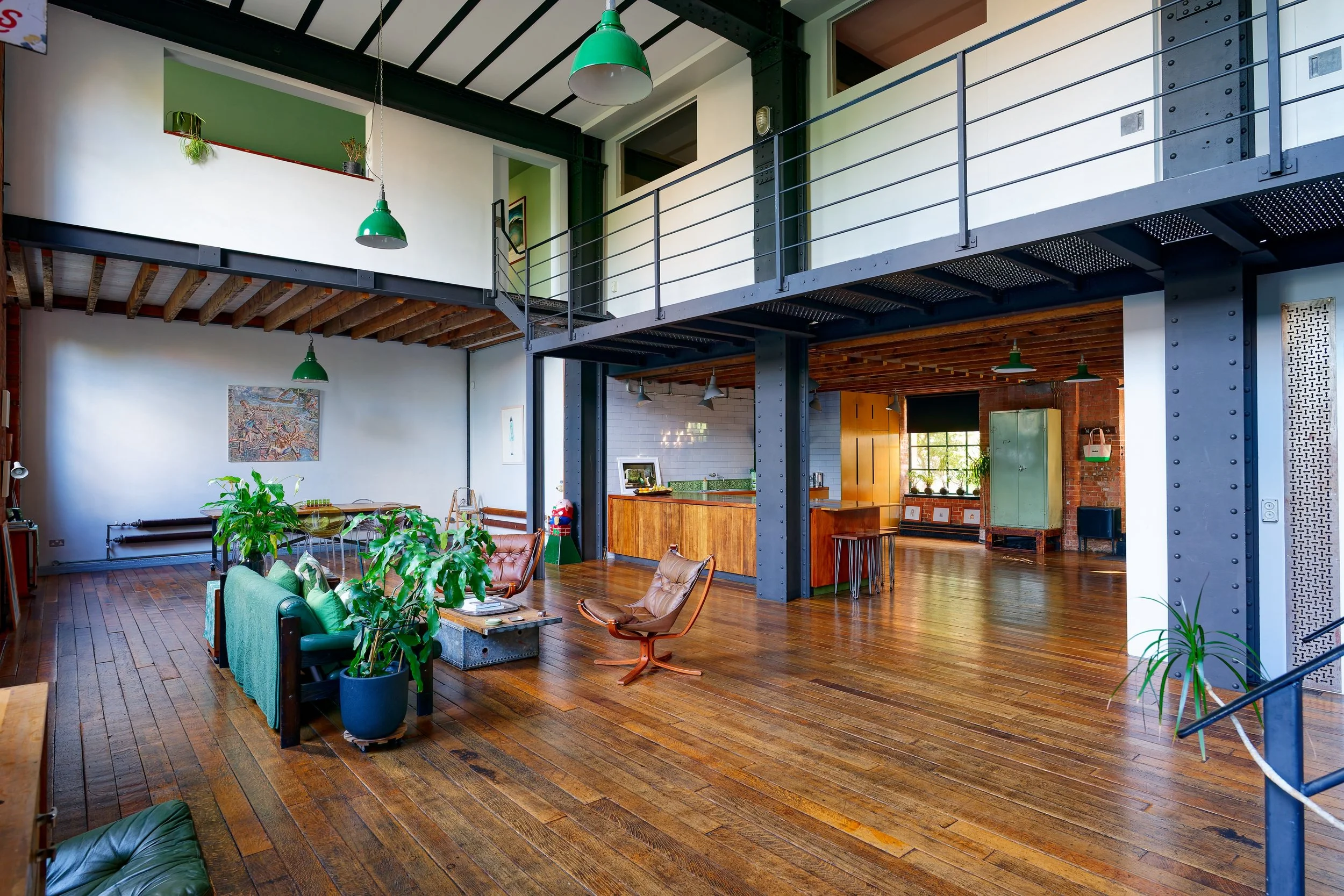 Spacious loft-style living room with wooden floors, exposed beams, a balcony, modern furniture, indoor plants, and large windows letting in natural light.