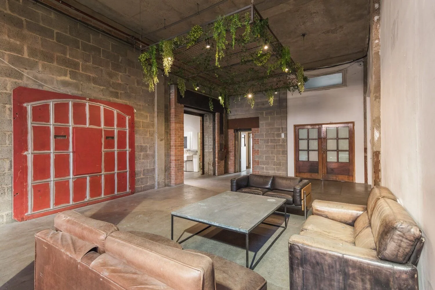 Industrial-style lounge with leather sofas, metal coffee table, exposed brick and concrete walls, and greenery hanging from a ceiling structure.