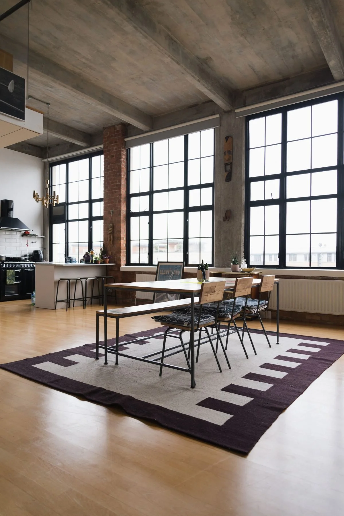Modern urban apartment dining area with wooden table, black metal chairs with patterned cushions, large windows, exposed brick and concrete ceiling, and a rug with geometric pattern.