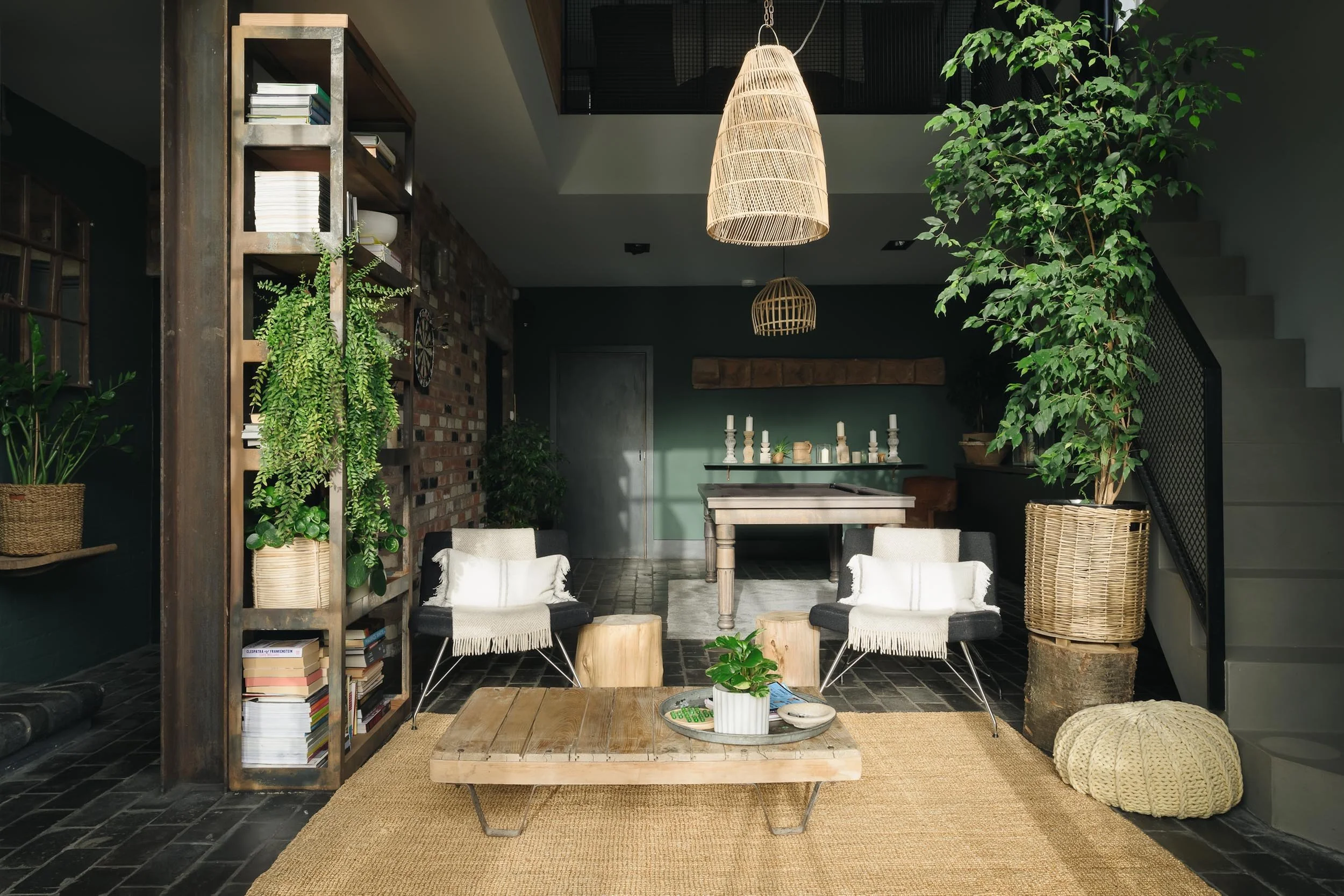 Interior of a modern living room with green walls, black tiled floor, a wooden bookshelf with plants, two black chairs with white cushions, a low wooden coffee table, large indoor plants, wicker lighting fixtures, and a staircase on the right.