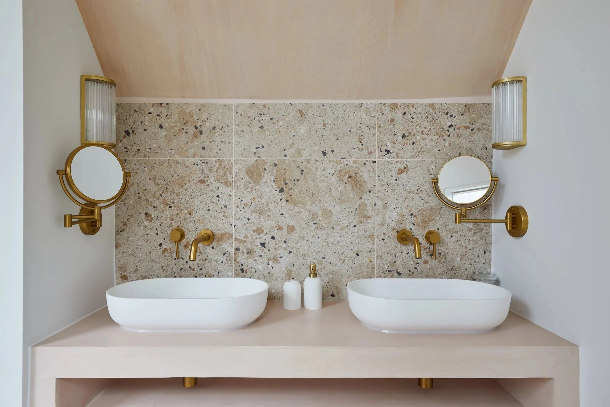 Bathroom vanity with two white vessel sinks, gold faucets, round mirrors with gold frames, wall-mounted light fixtures, and a terrazzo backsplash.
