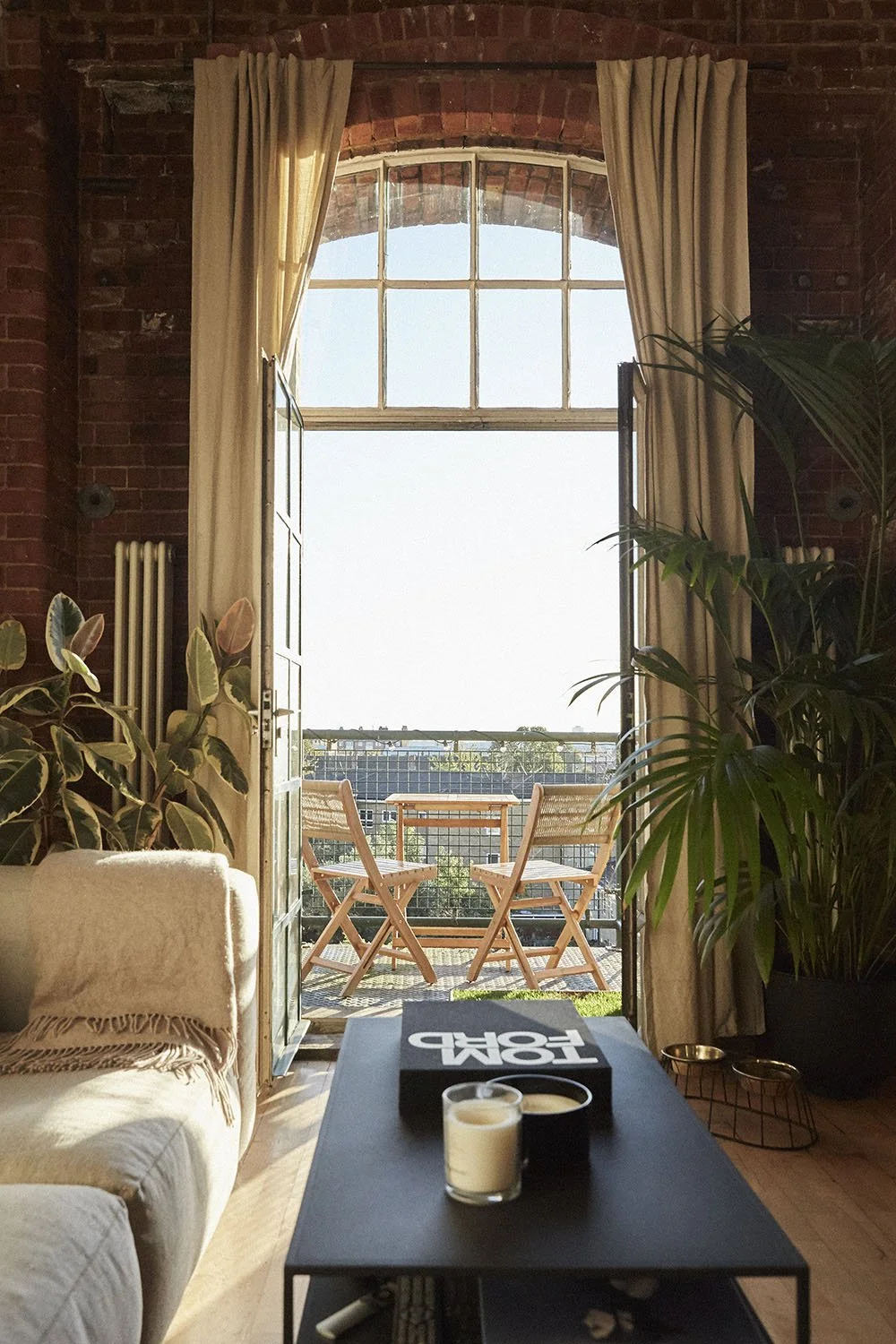 View from inside a living room through an open glass door to a balcony with wooden chairs and a table, surrounded by plants and brick walls.