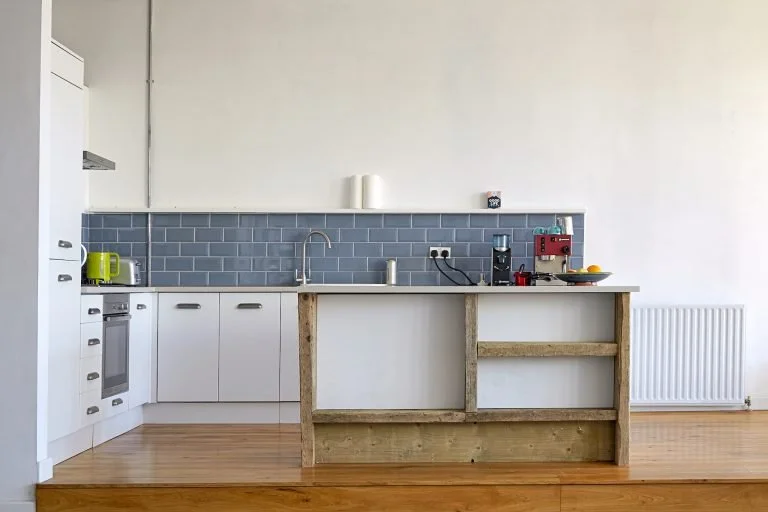 Minimalist kitchen with white cabinets, blue subway tile backsplash, wooden floor, and a makeshift island with a wooden frame.