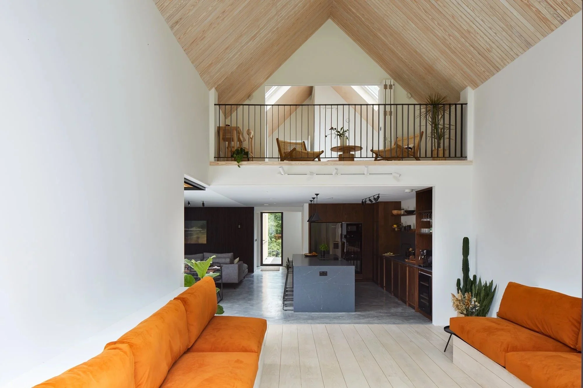 Open-concept living space with white walls, light wood floors, and vaulted ceiling. Orange sofas at the foreground, view into kitchen with dark wood cabinetry and an island, and a loft with seating area and plants above.