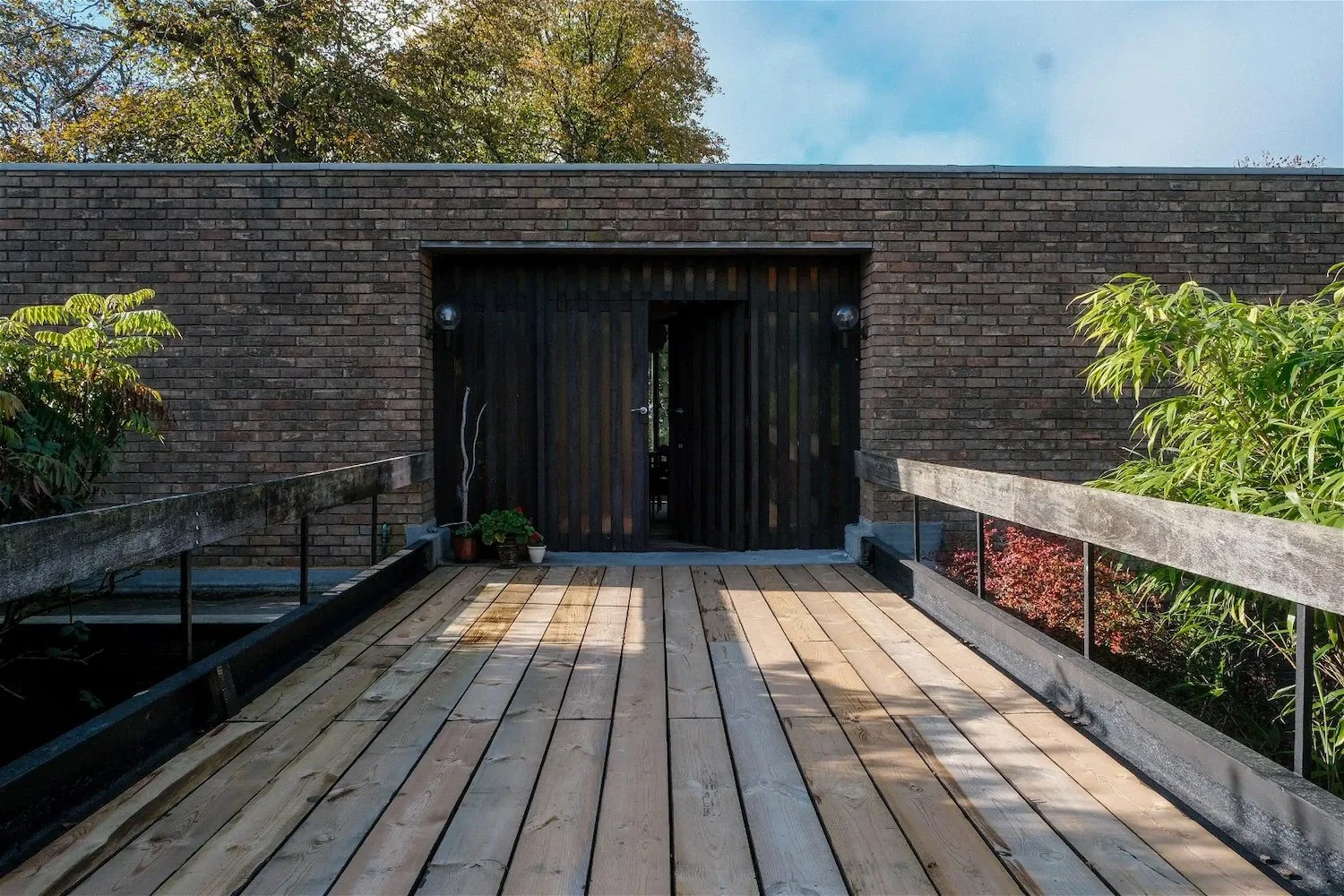 Wooden deck leading to a dark wooden door framed by brick wall, with plants on either side and a blue sky above.