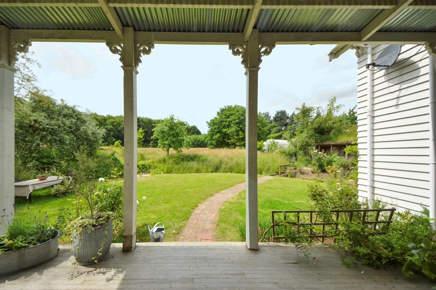 View from a porch looking out onto a green backyard with a brick path, trees, and a field in the distance. There are potted plants on the porch and a white house with siding on the right.