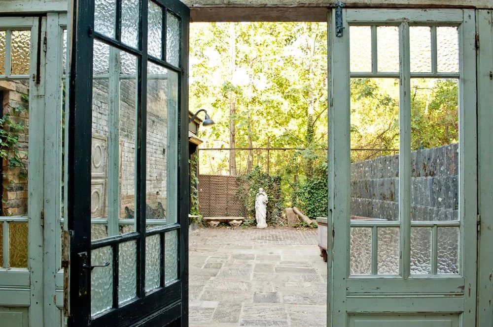 View through open glass doors to a patio with stone flooring, greenery, and a stone wall.