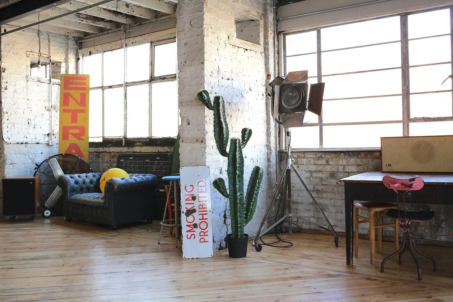 Interior of an industrial-style room with exposed brick walls and large windows. There is a black sofa with a yellow ball on it, a vintage fan, a green potted cactus next to a 'Smoking Prohibited' sign, and a large speaker on a stand. There's a table