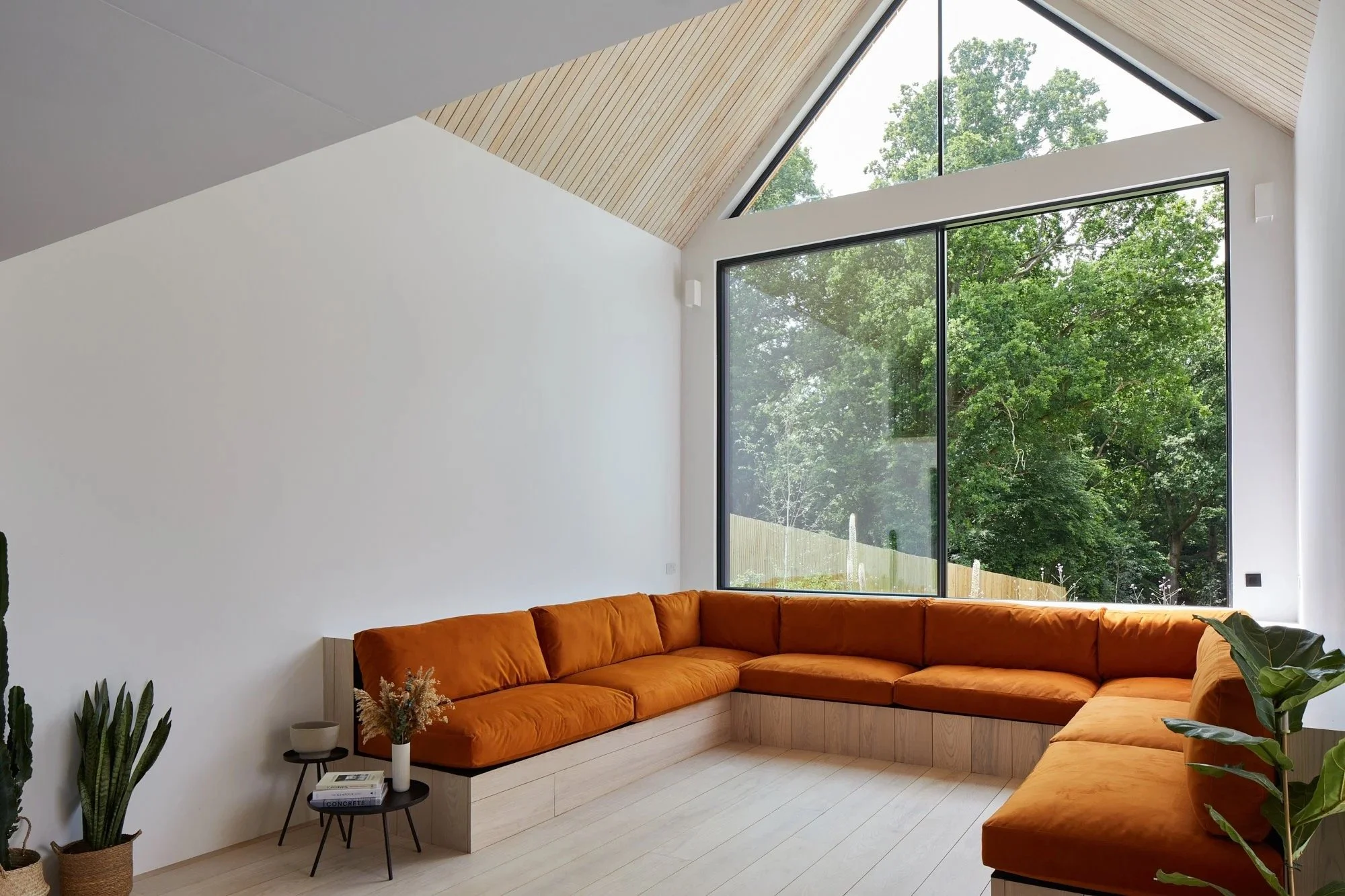 Modern living room with white walls, large window showing greenery outside, and an orange sectional sofa with wooden built-in base.