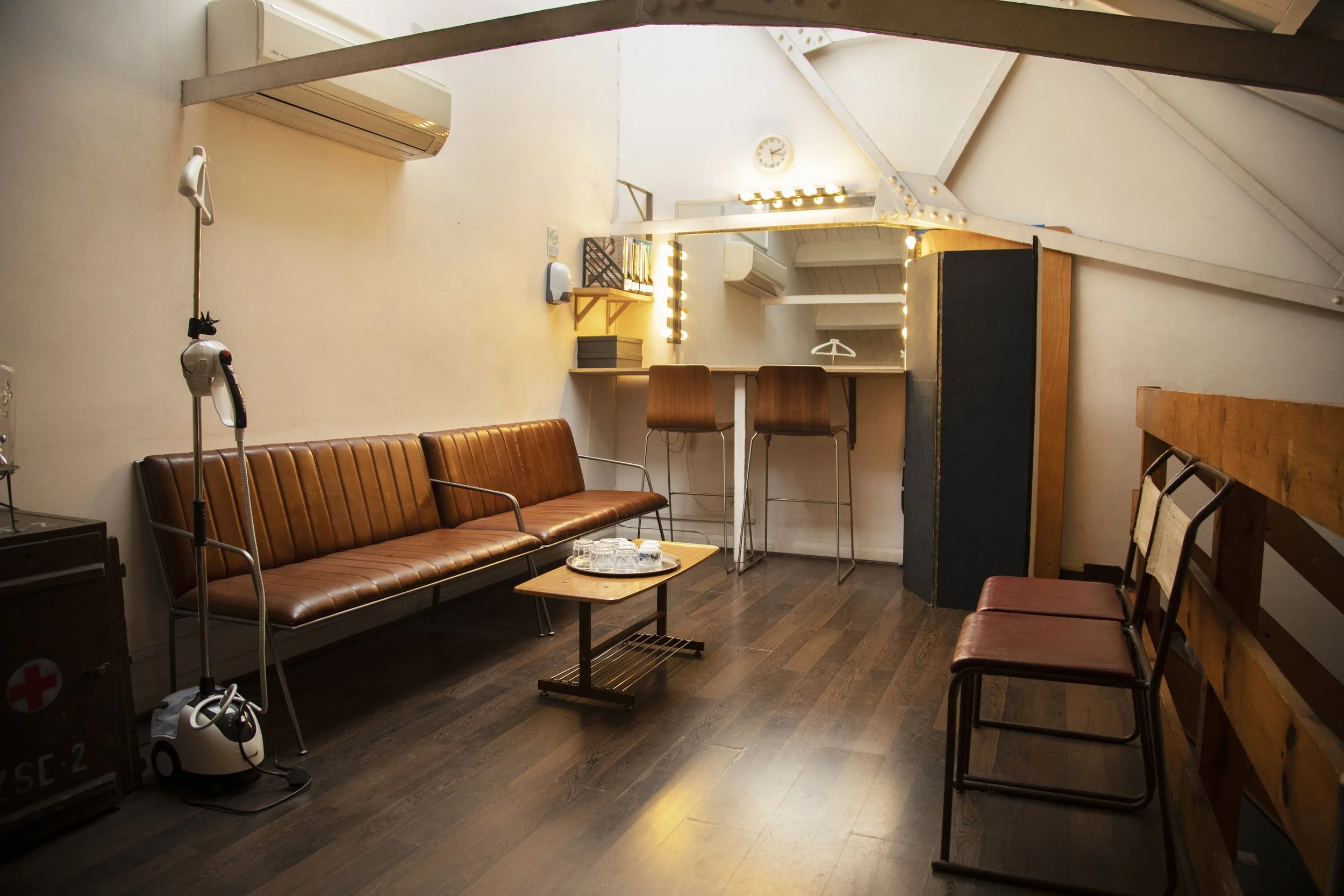 A cozy room with brown leather sofas, a small wooden table with glasses, a bar-style counter with two chairs, and various decorations including a clock, lights, books, and an IV stand.