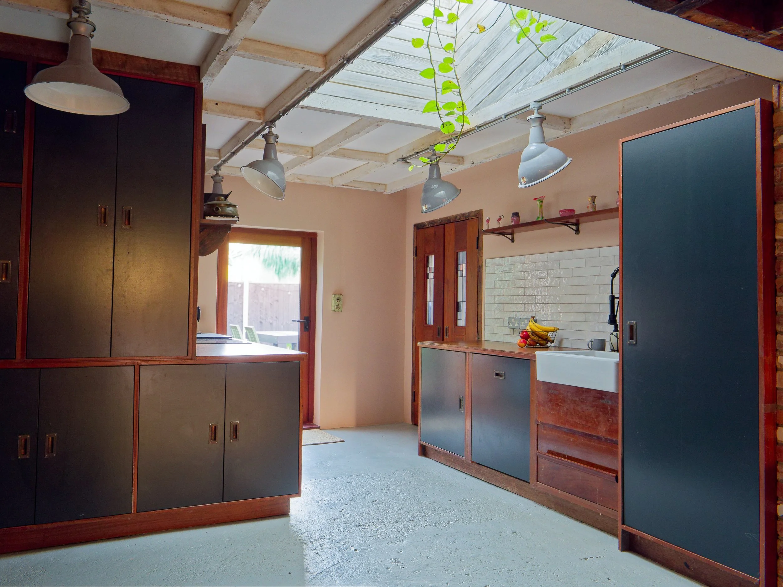 A rustic kitchen with dark blue cabinets, wooden accents, a white farmhouse sink, hanging industrial-style light fixtures, a skylight with a hanging plant, and a door leading outside.