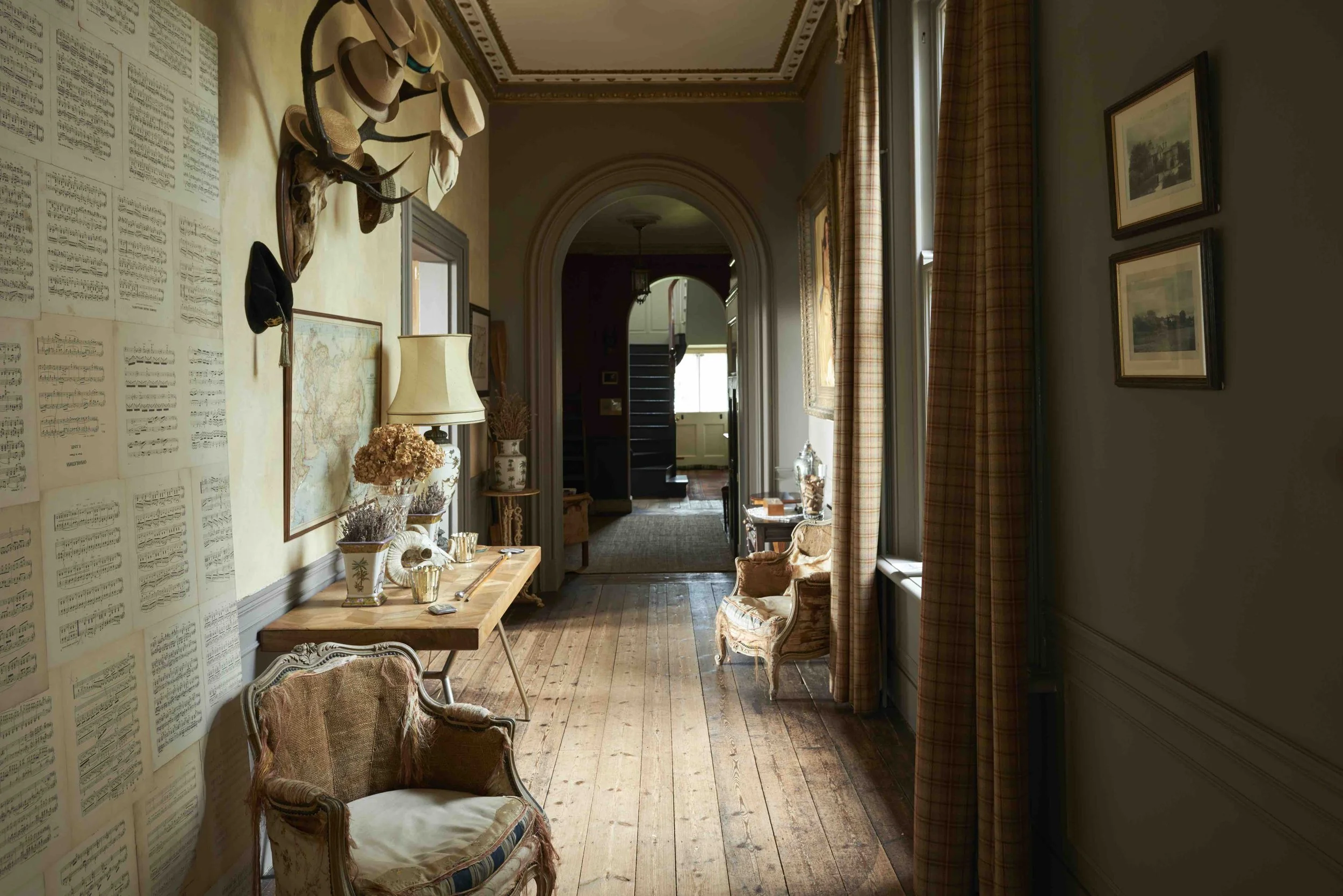 Interior of a vintage-style hallway with wood flooring, framed artwork, hats hanging on the wall, a side table with dried flowers, a lamp, and armchairs near the windows with plaid curtains.