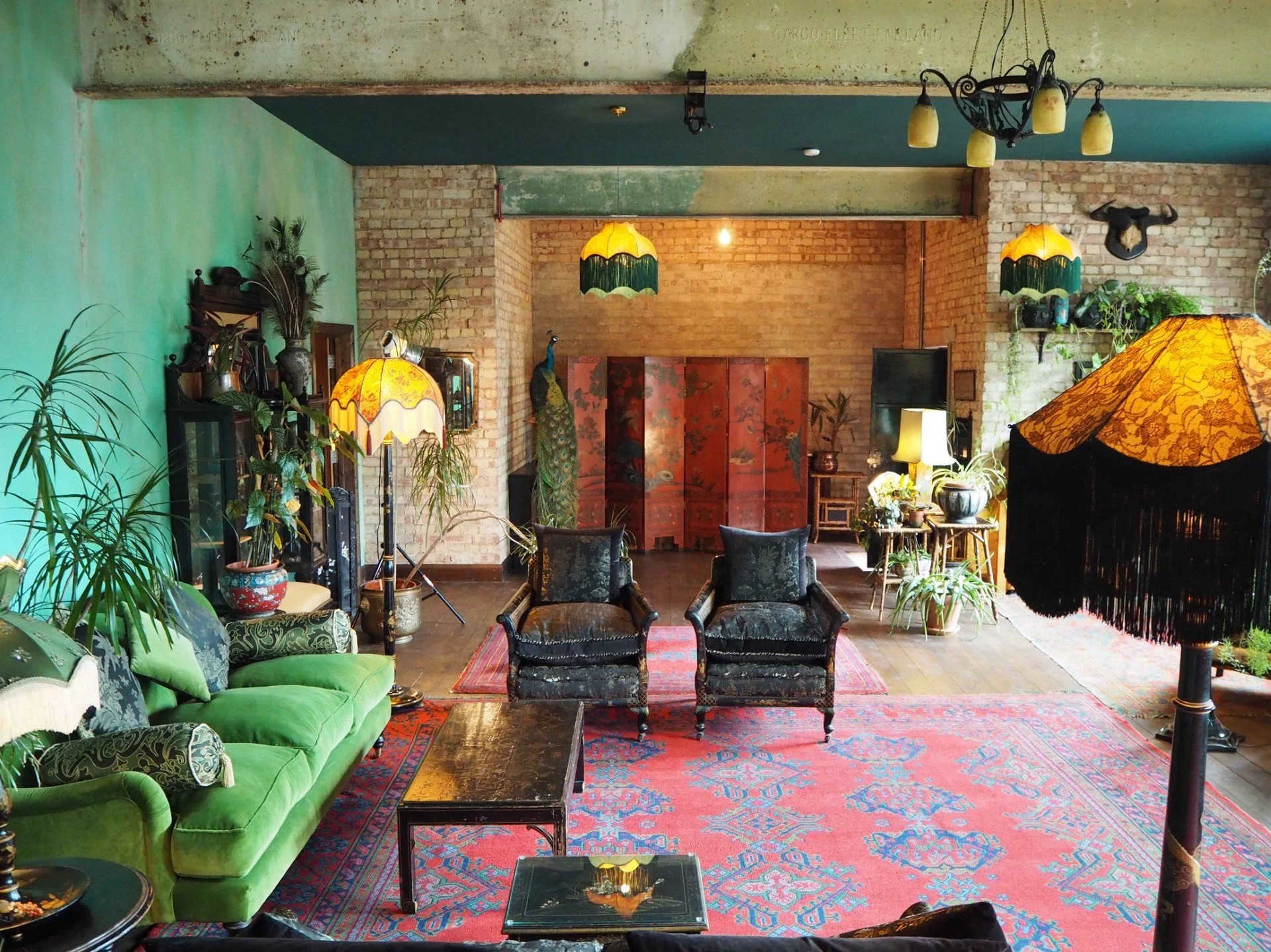 A vintage living room with green sofa, black velvet armchairs, wooden side tables, and decorative lamps. Yellow lampshades, indoor plants, brick walls, and colorful rugs add to the eclectic decor.