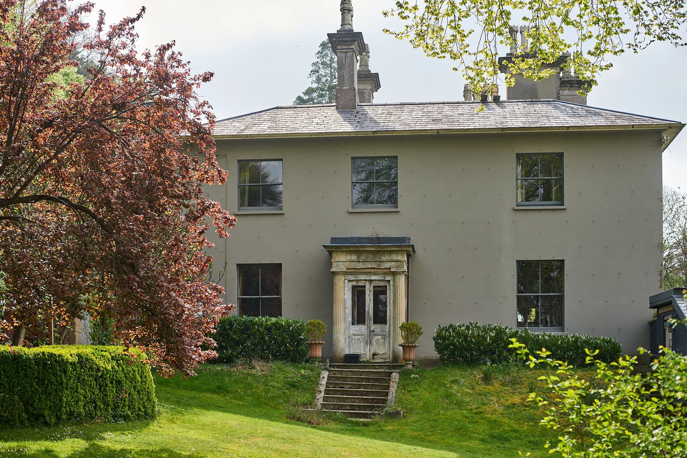 Front view of a three-story, light-colored house with a decorative stone entryway, surrounded by lush green trees and bushes.
