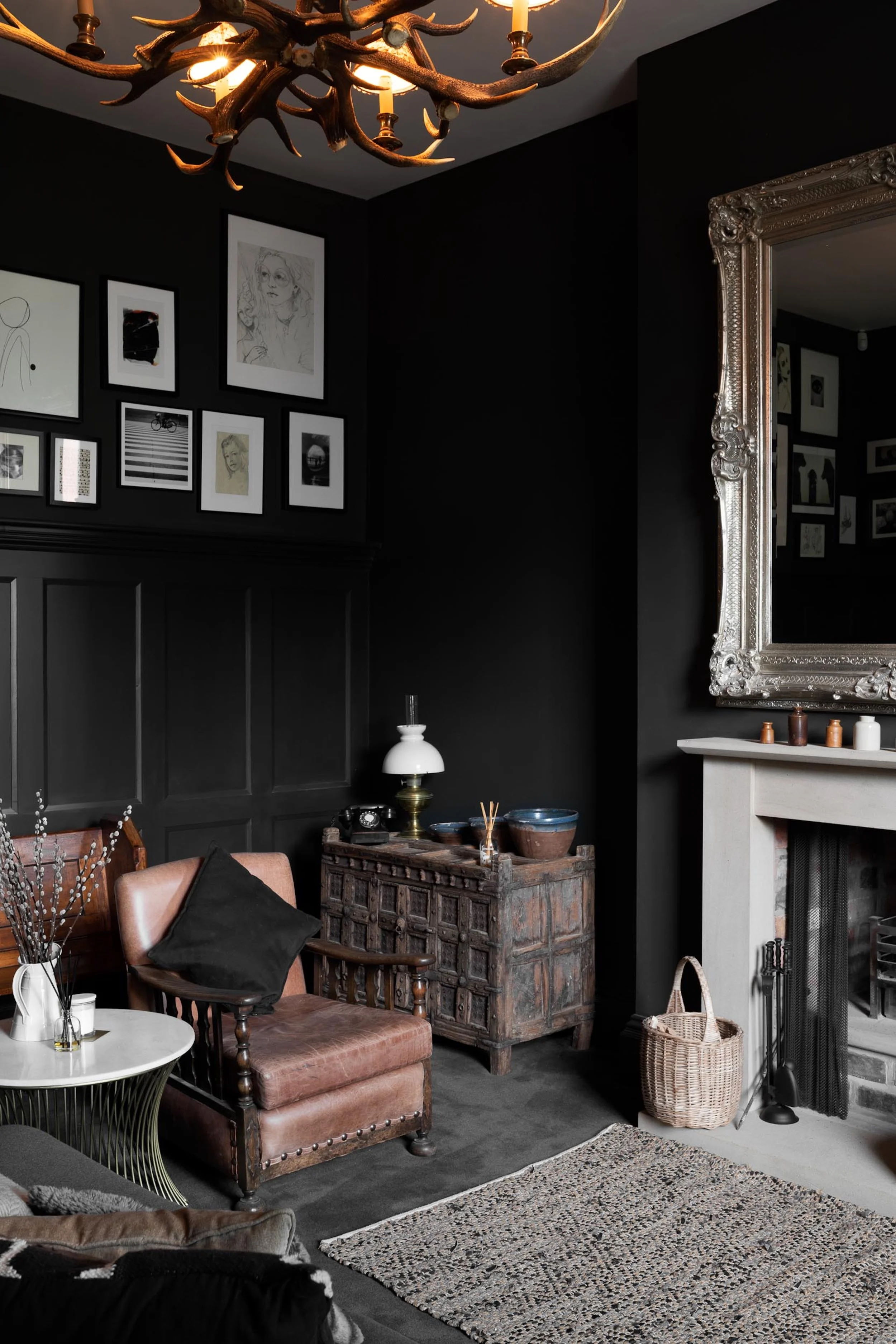A cozy living room corner with black painted walls, a vintage brown leather chair with a black pillow, a white side table with vases, a decorative vintage wooden cabinet with bowls and a lamp, a large ornate mirror above a white fireplace mantel, a w