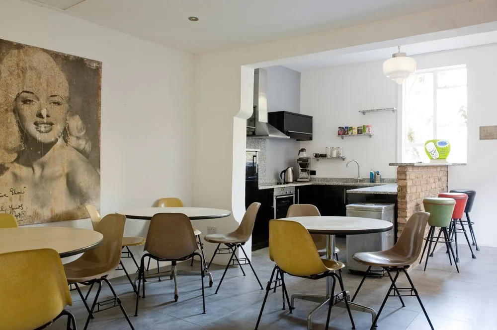 A bright, modern dining area with round tables and colorful chairs, adjacent to a kitchen with black cabinets, stainless steel appliances, and a small bar area with stools. A large black-and-white portrait of Marilyn Monroe hangs on the wall.