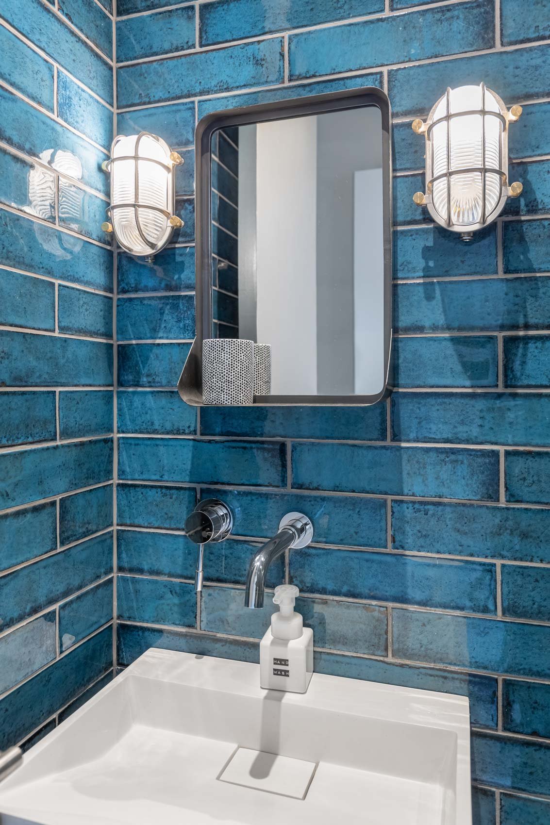 A bathroom with blue tiled walls, a rectangular mirror, two wall sconces, a white sink with a soap dispenser, and a wall-mounted faucet.