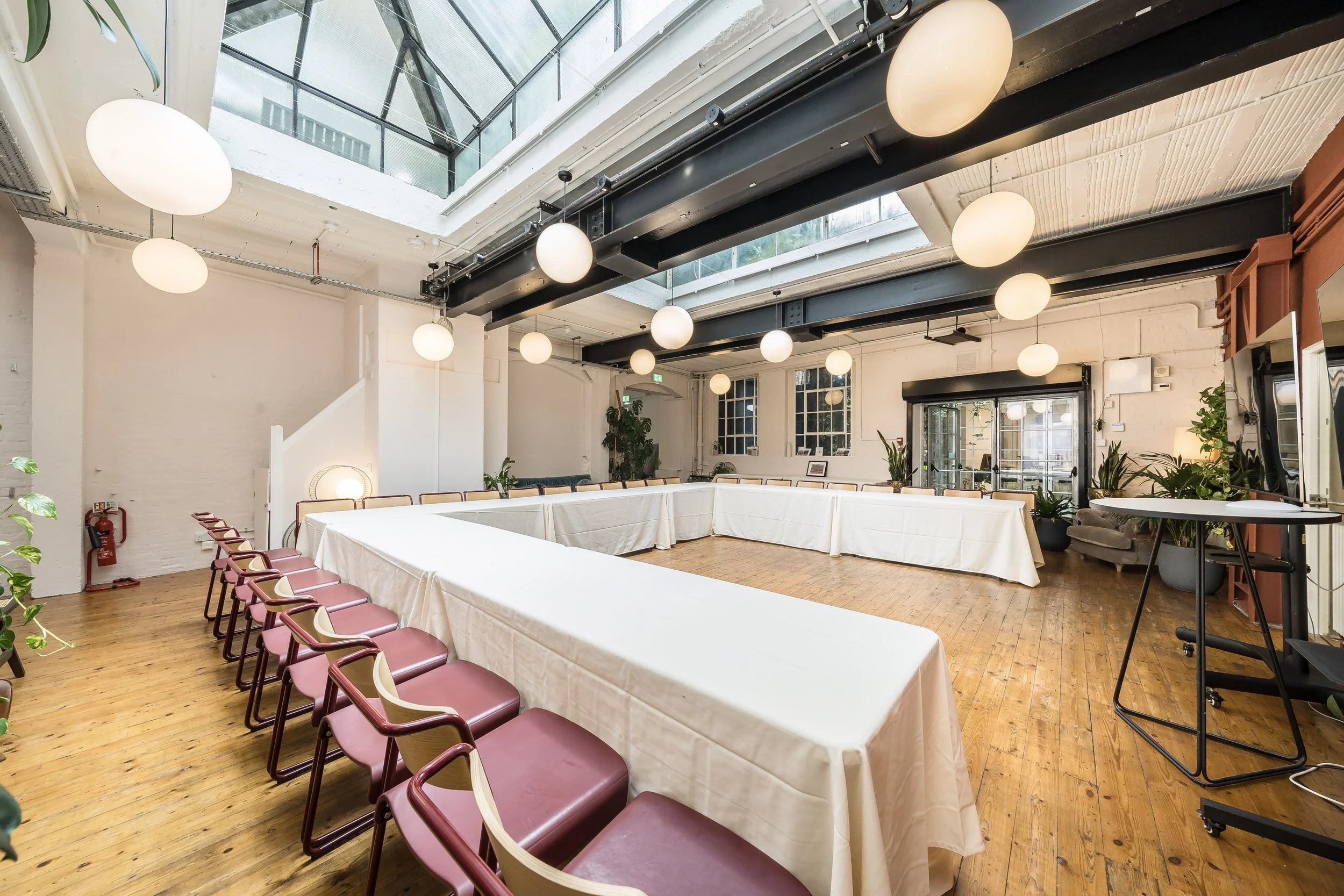 A spacious conference or meeting room with natural light coming through a glass ceiling. The room has a U-shaped table arrangement with white tablecloths, surrounded by pink chairs with wooden armrests. The floor is hardwood, and the room features wh