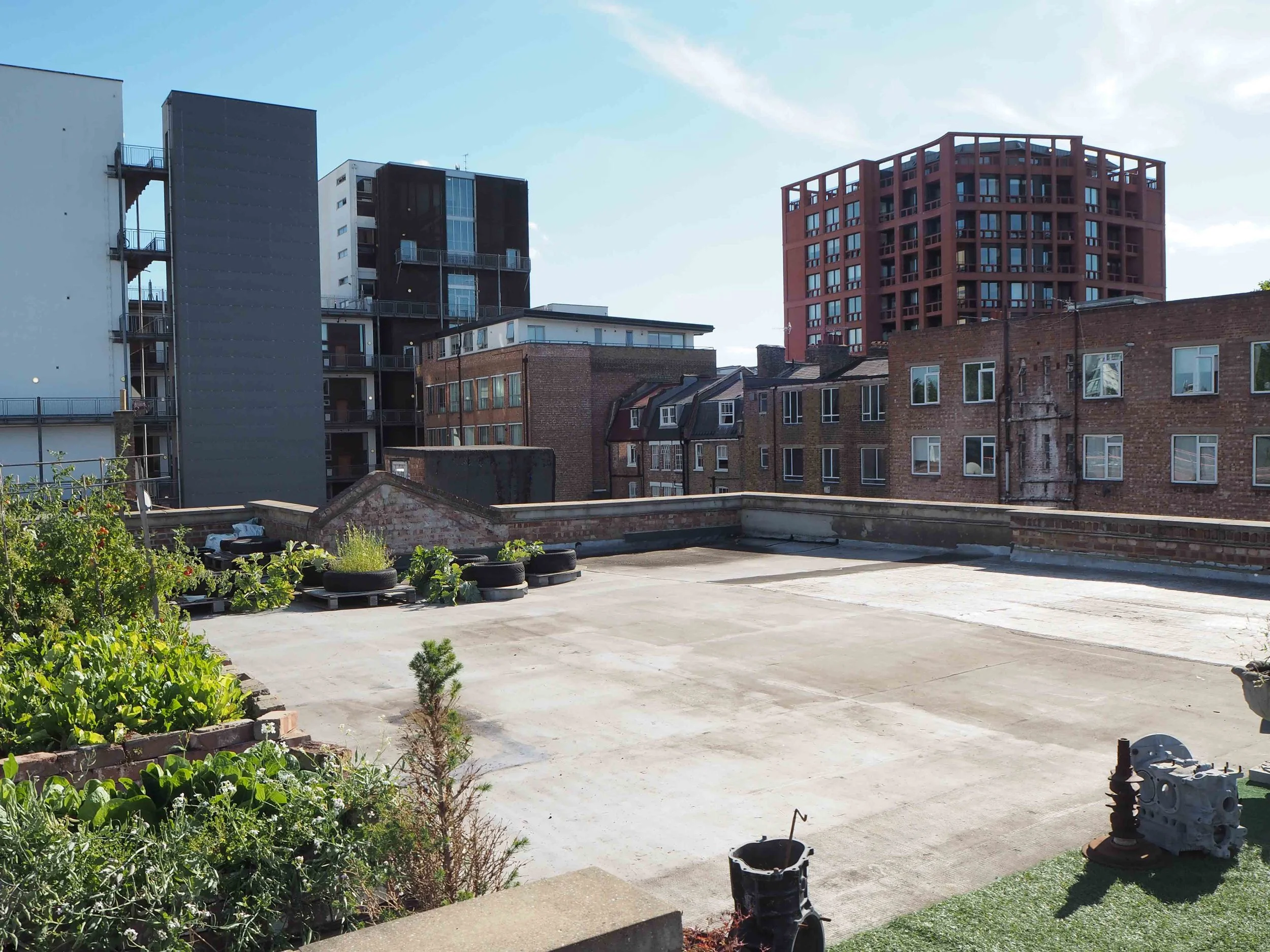 Rooftop in an urban area with garden patches, tires, and construction materials, surrounded by multistory buildings and a partly cloudy sky.