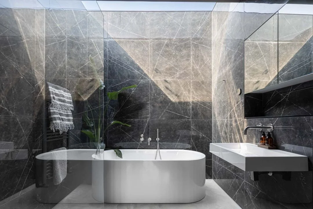 Modern bathroom with a freestanding white bathtub, dark marble walls, a small white sink with a black shelf below, a gray towel on a black towel rack, a green plant, and a skylight ceiling.