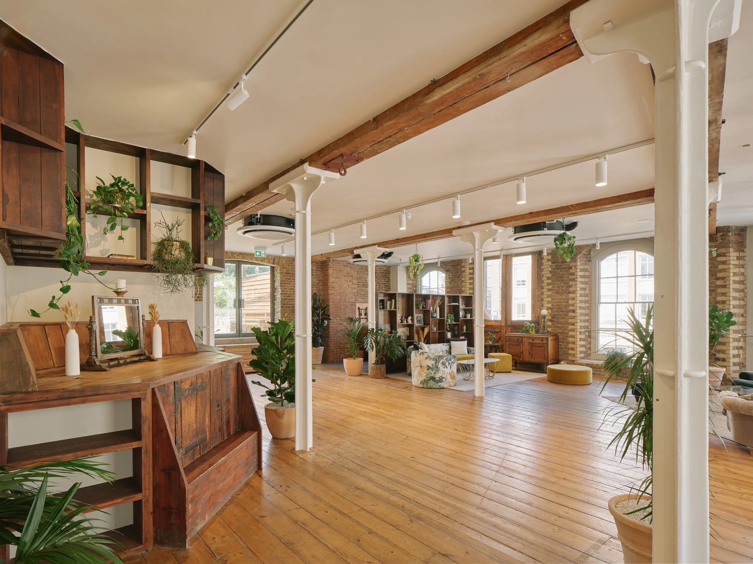 Open living room with wooden floors, brick walls, large windows, and various potted plants.