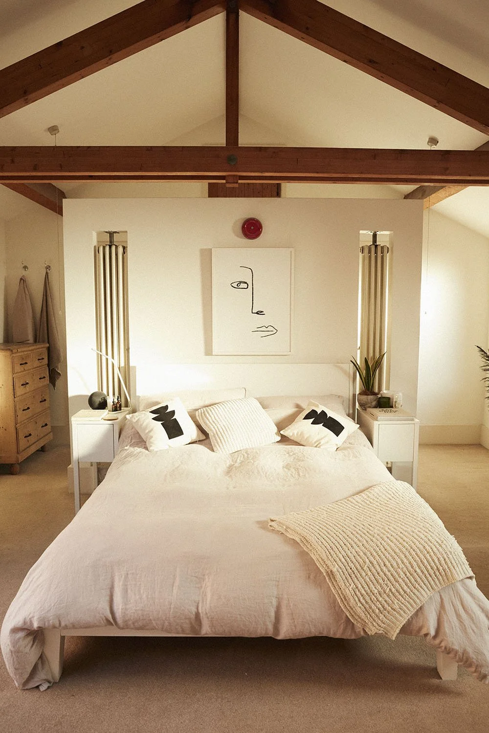 A cozy bedroom with a large bed covered in light pink bedding, two black and white accent pillows, a cream knitted blanket, and a central pillow. Small white nightstands with lamps and decorative items are on each side of the bed, against a white wal