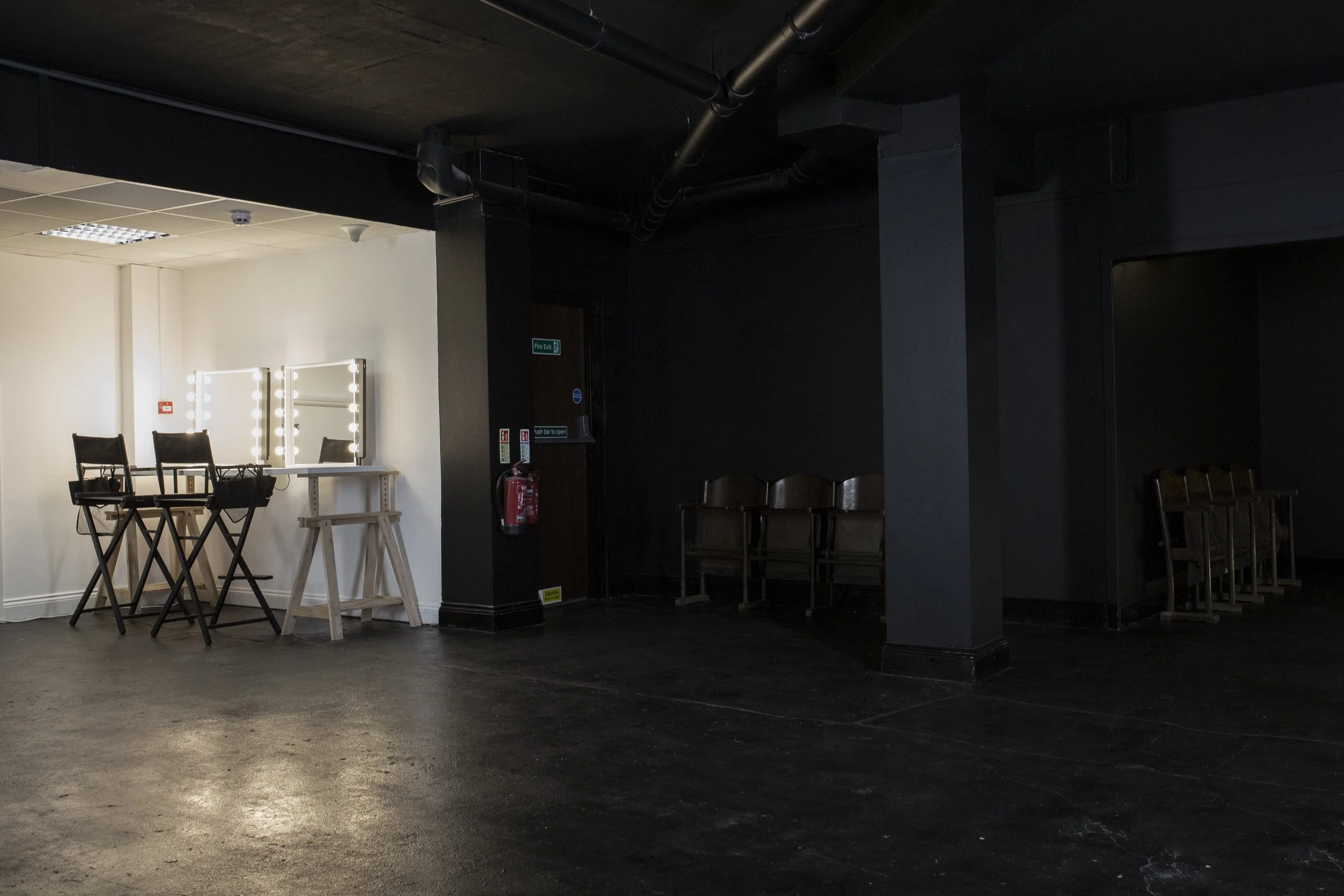 Empty backstage or dressing room area with two illuminated mirrors and makeup chairs on the left, and rows of chairs in the dark area on the right, with black walls and overhead pipes.