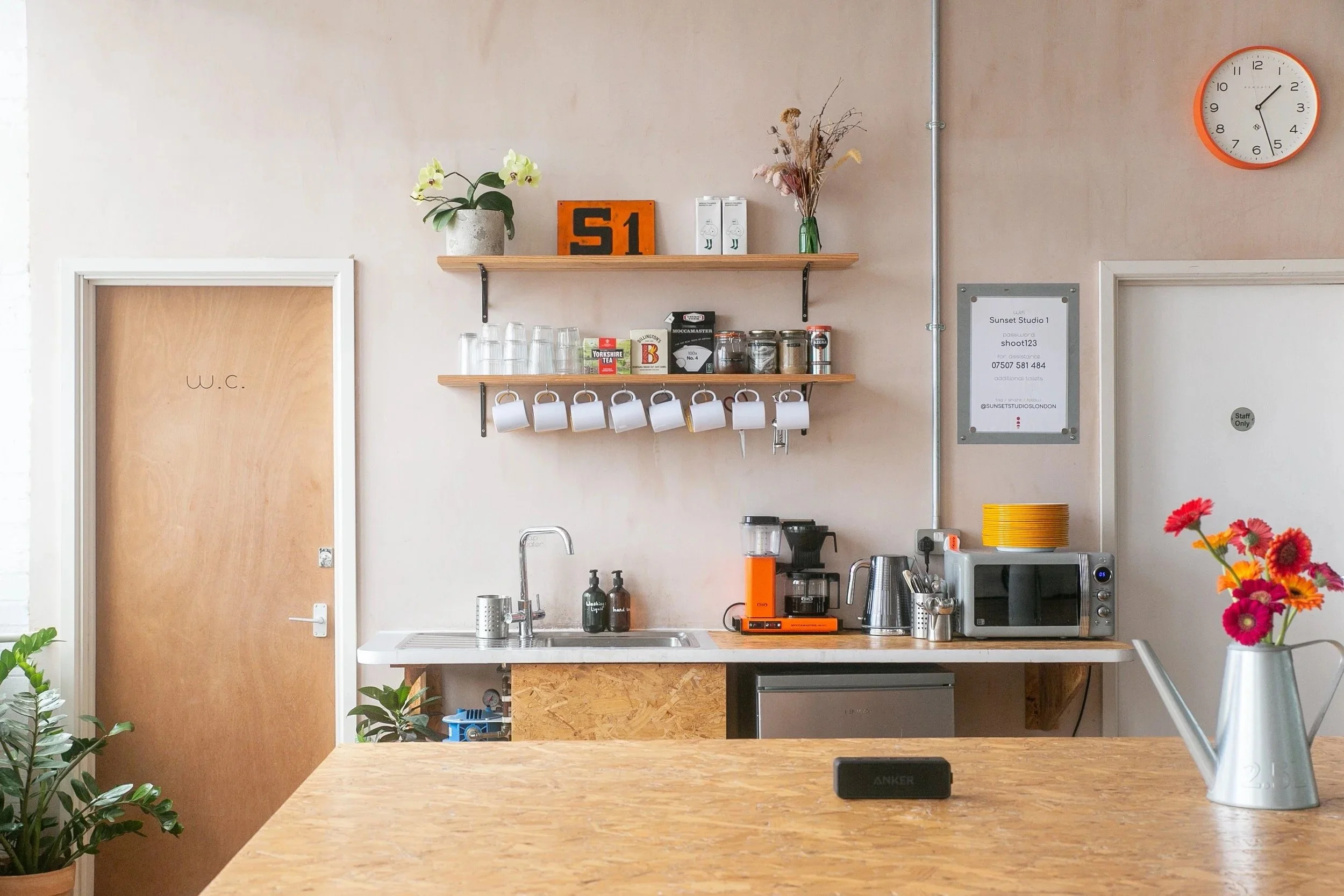 A modern kitchen with wooden shelves holding glasses, cups, and jars, a coffee maker, microwave, and other appliances on the counter, and a vase with pink and orange flowers on a wooden table in the foreground.