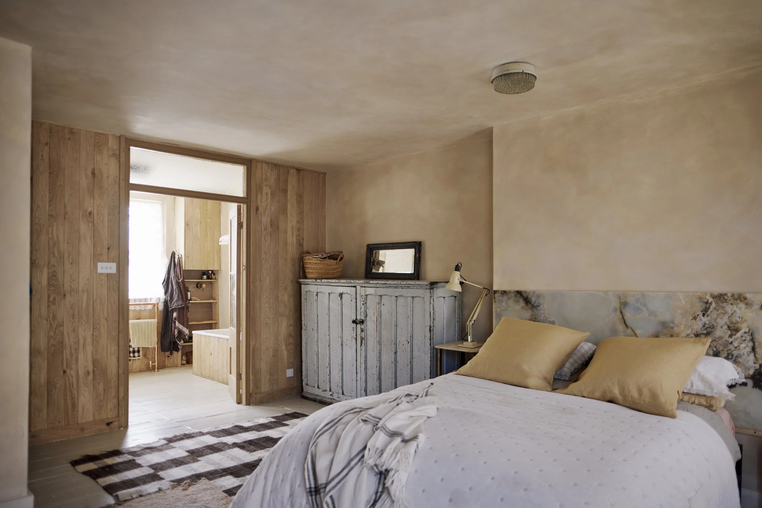 Cozy bedroom with a bed featuring yellow and white pillows, a rustic white wardrobe, wooden walls, and an adjoining bathroom visible through an open door.
