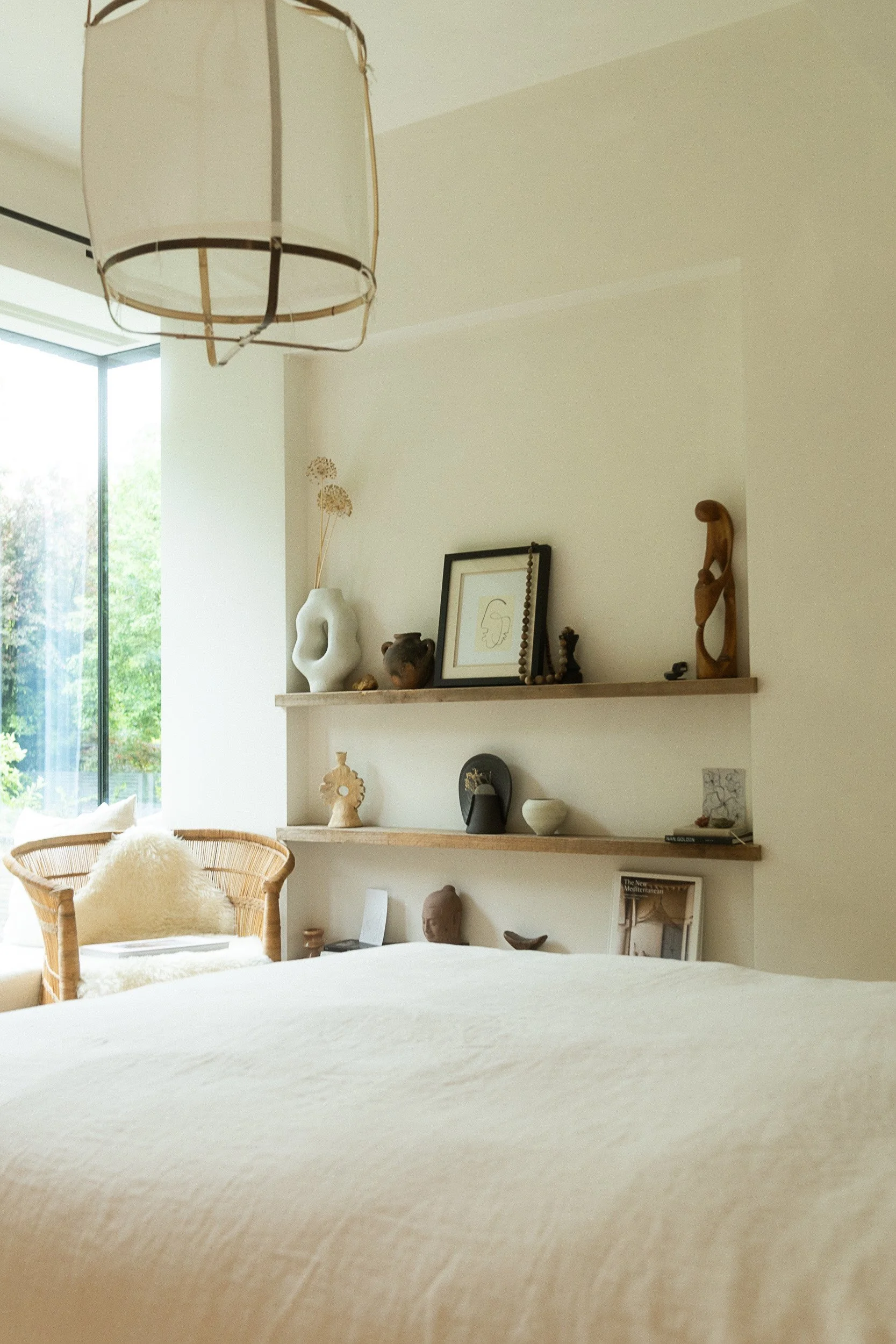 A bedroom corner with a bed covered in a light-colored bedspread, a rattan chair with a fluffy pillow, a large window letting in natural light, and floating shelves on the wall displaying decorative objects like vases, sculptures, and framed artwork.