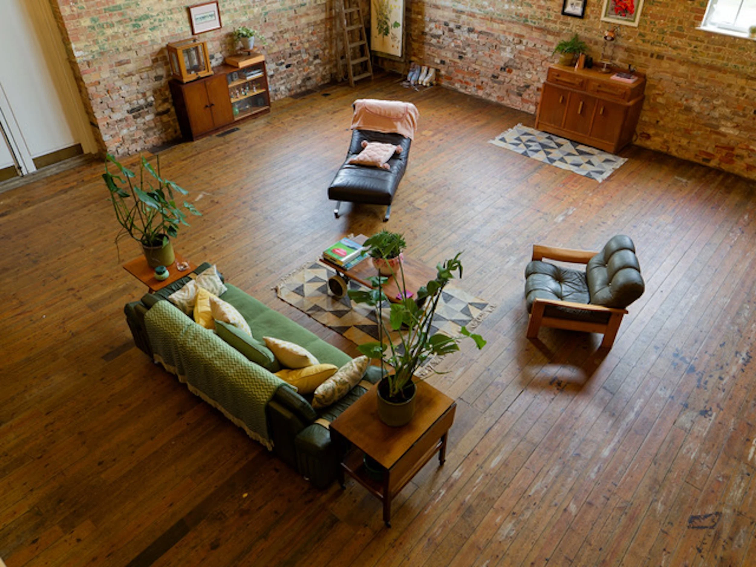 Living room with exposed brick walls, wooden floor, green sofa with yellow and beige pillows, gray armchair, black chaise lounge, and wooden furniture including a cabinet and side table with plants.