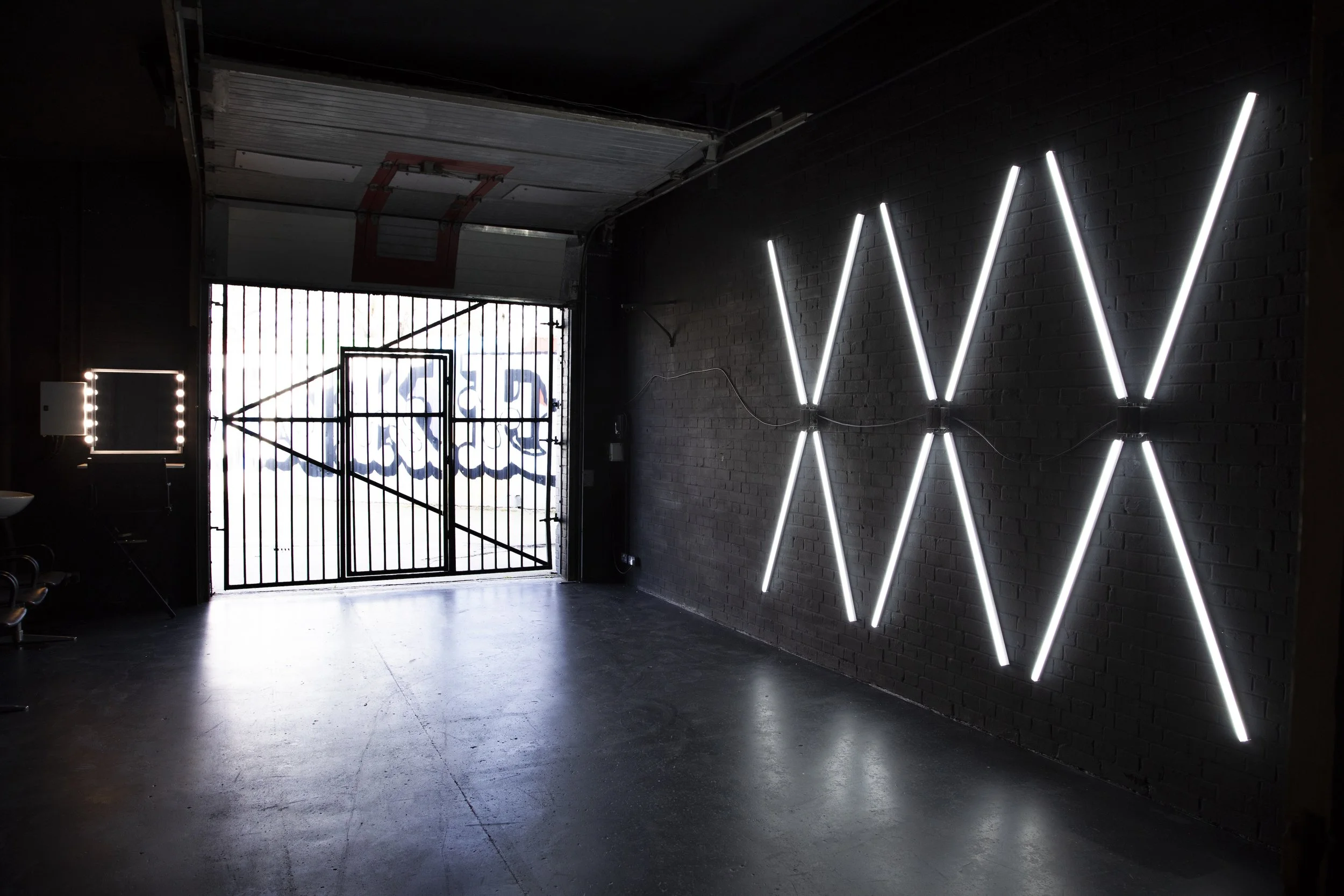 Interior of a modern art gallery with black brick walls, illuminated white neon lights arranged in an X pattern on the right wall, and a black metal gate with a geometric design and a graffiti-style artwork visible outside through the open garage doo